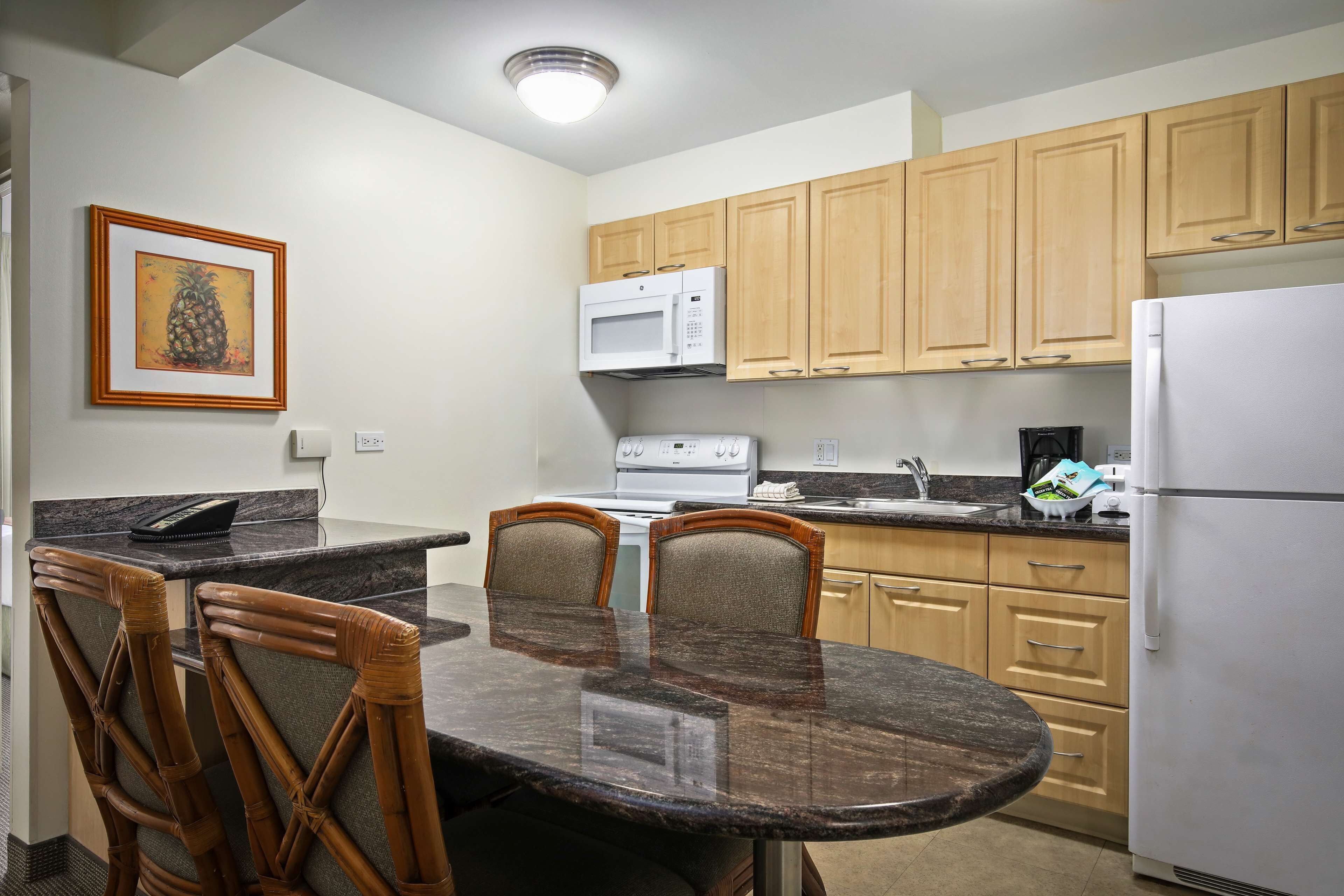 A kitchen with wooden cabinets, a microwave, a stove, and a refrigerator. There is a dining table with chairs and a framed picture on the wall.