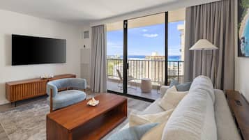 A modern living room with a large flat-screen TV, a sofa, a coffee table, and a view of the ocean through large sliding doors.