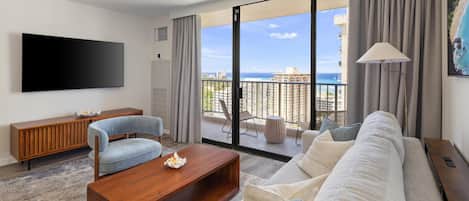 A modern living room with a large flat-screen TV, a sofa, a coffee table, and a view of the ocean through large sliding doors.