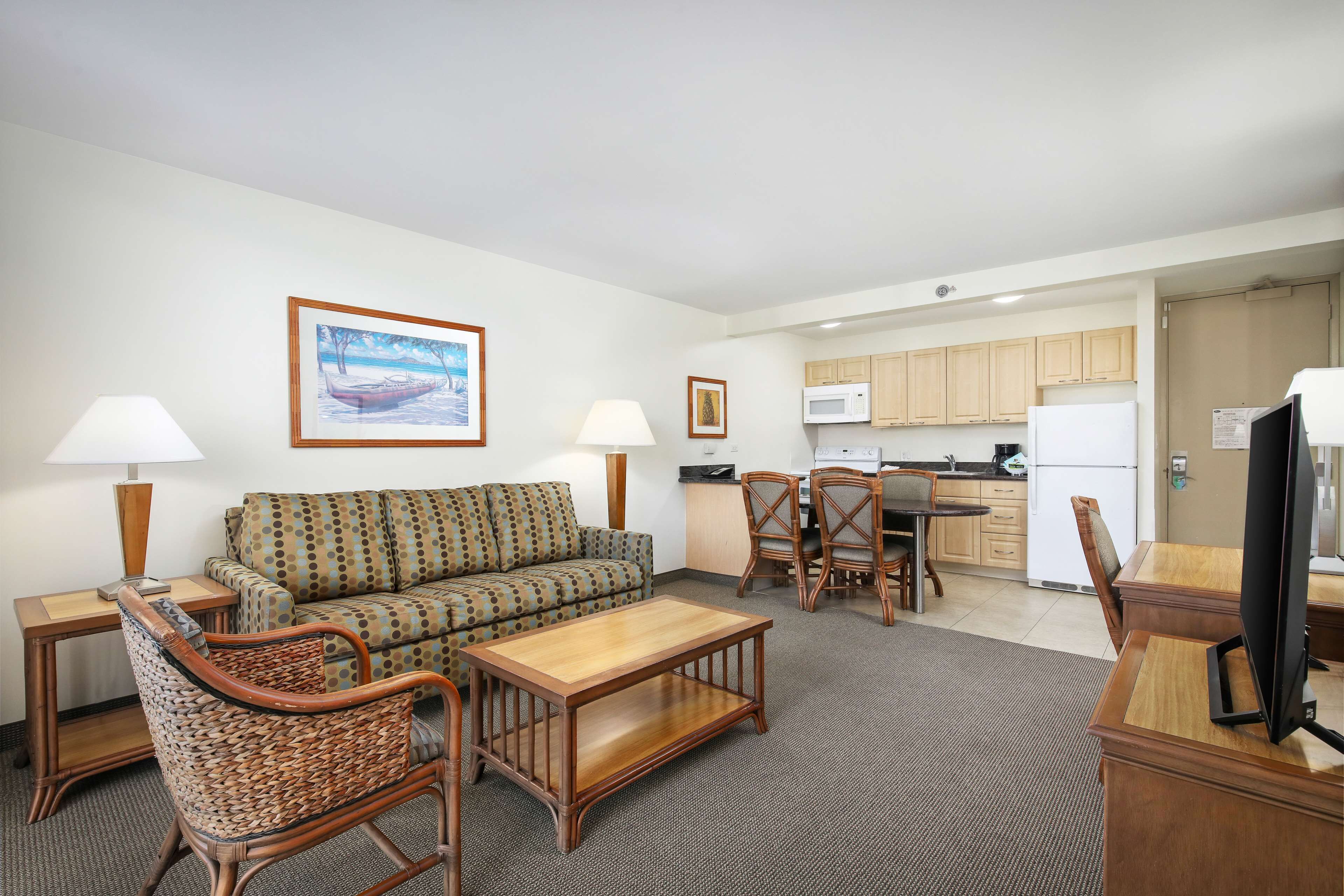 A living room with a sofa, chairs, a coffee table, and a TV. There is a kitchen area with cabinets and appliances.