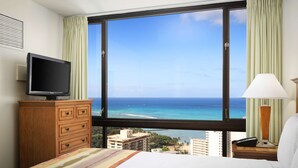 A hotel room with a bed, a television on a wooden dresser, a nightstand with a lamp, and a large window offering a view of the ocean and buildings. - Aston Waikiki Sunset (Honolulu)