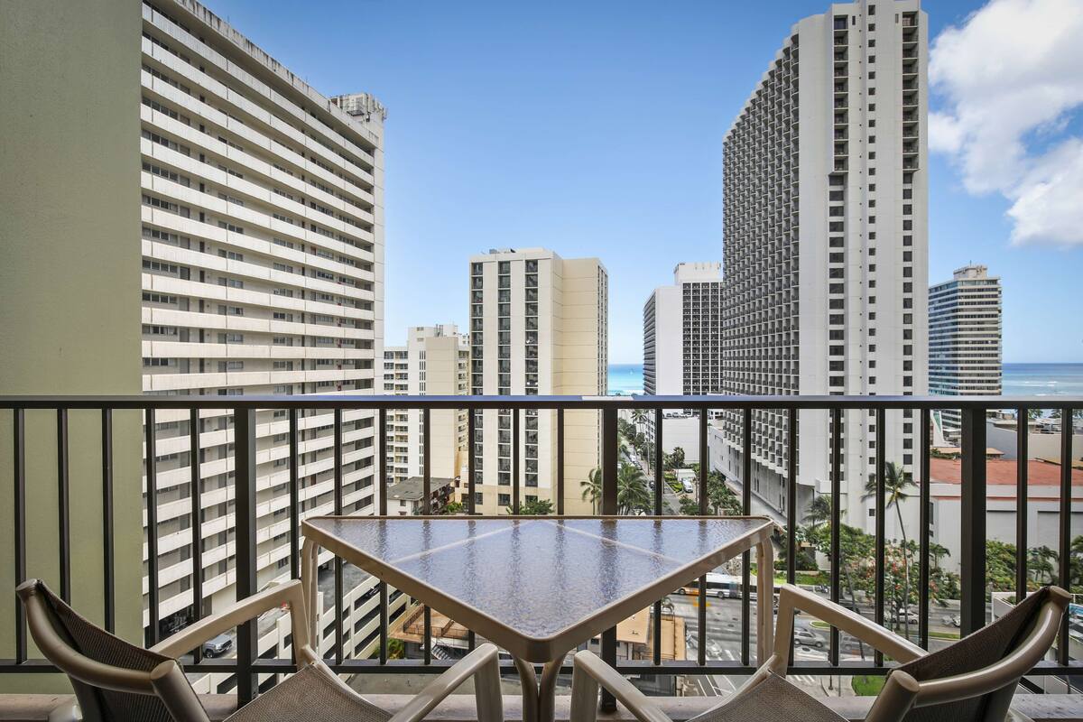 A balcony with a table and chairs, overlooking a cityscape with tall buildings and a clear sky.