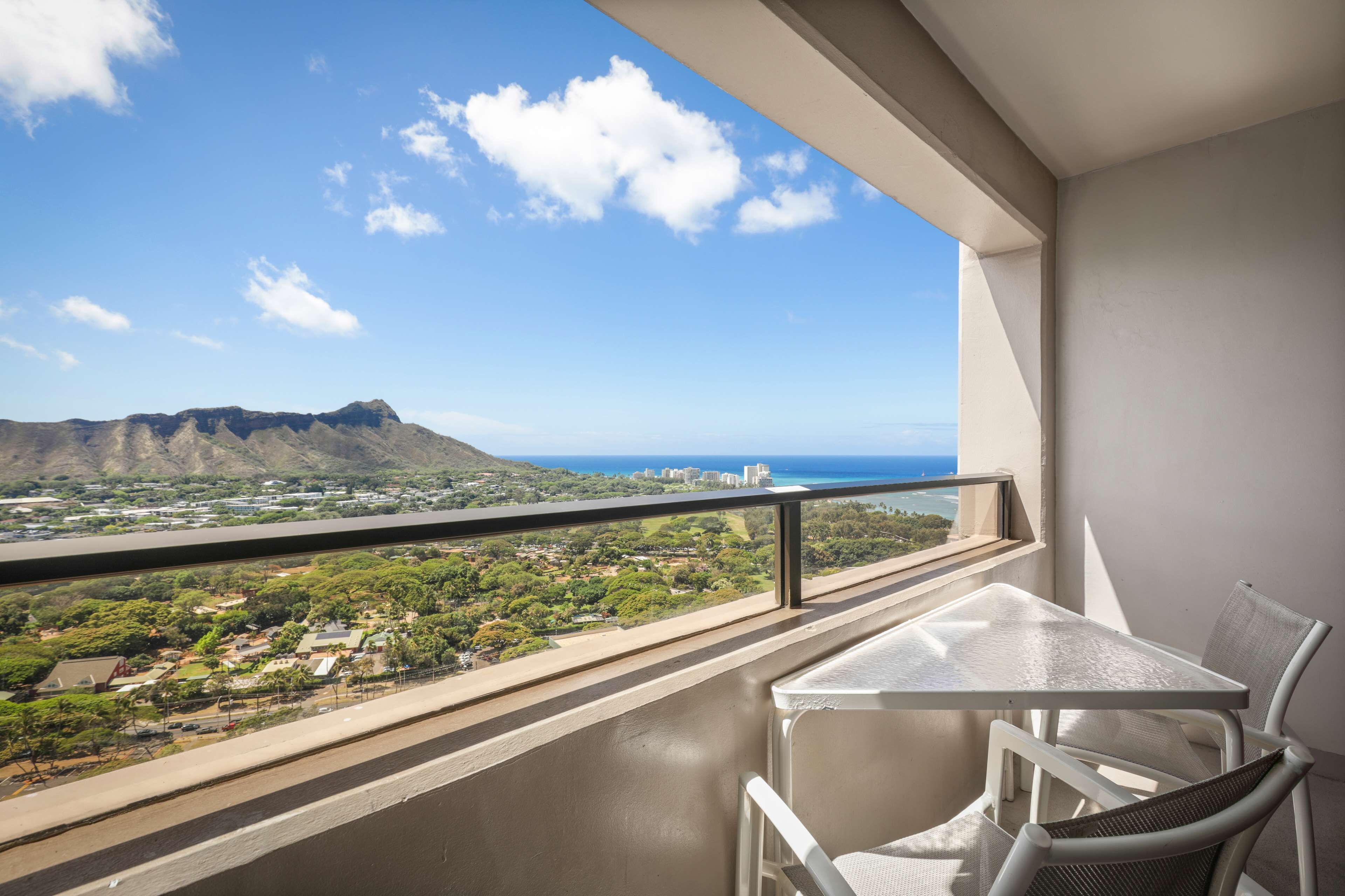 A balcony with a table and chairs, overlooking a coastal landscape with mountains and buildings.