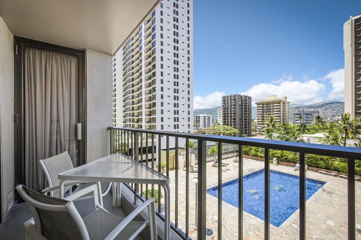 A balcony with a table and chairs, overlooking a swimming pool and a cityscape.