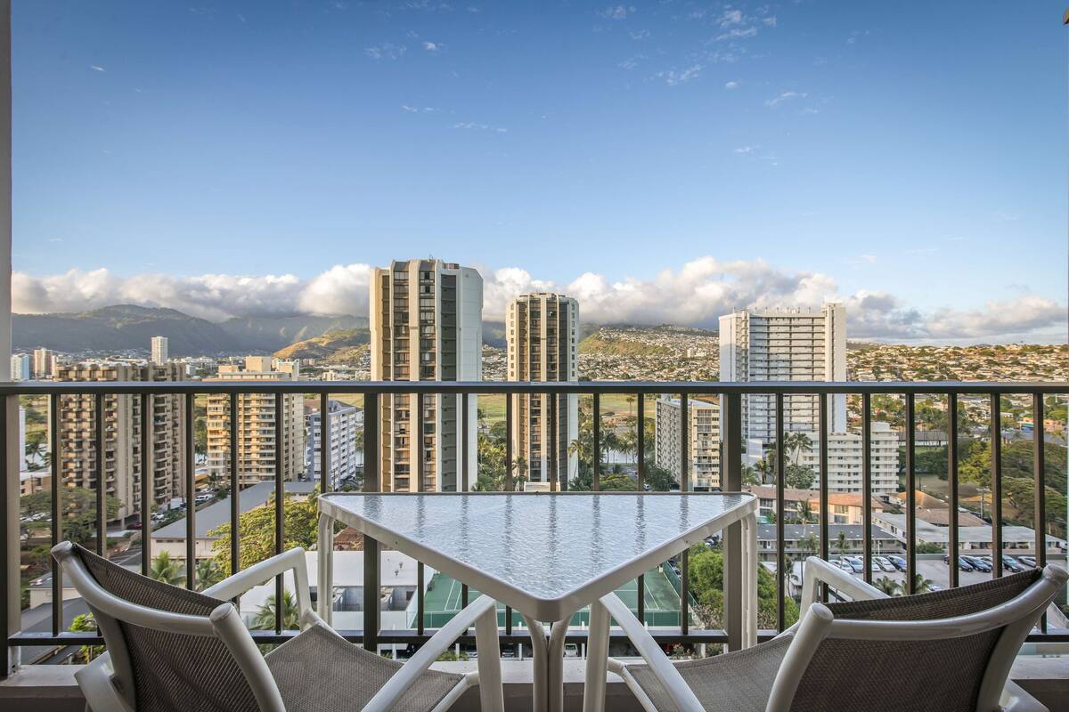 A balcony with a table and chairs, offering a view of tall buildings and mountains.