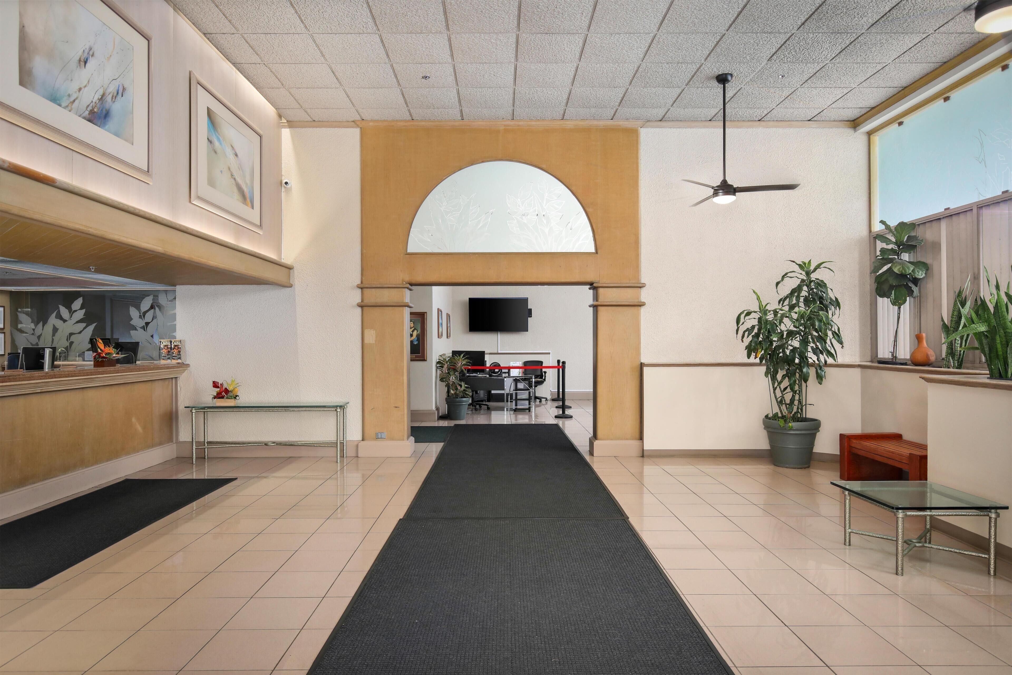 Un vestíbulo de hotel con mostrador de recepción, una alfombra negra que conduce a una sala de espera, un televisor y plantas en macetas.