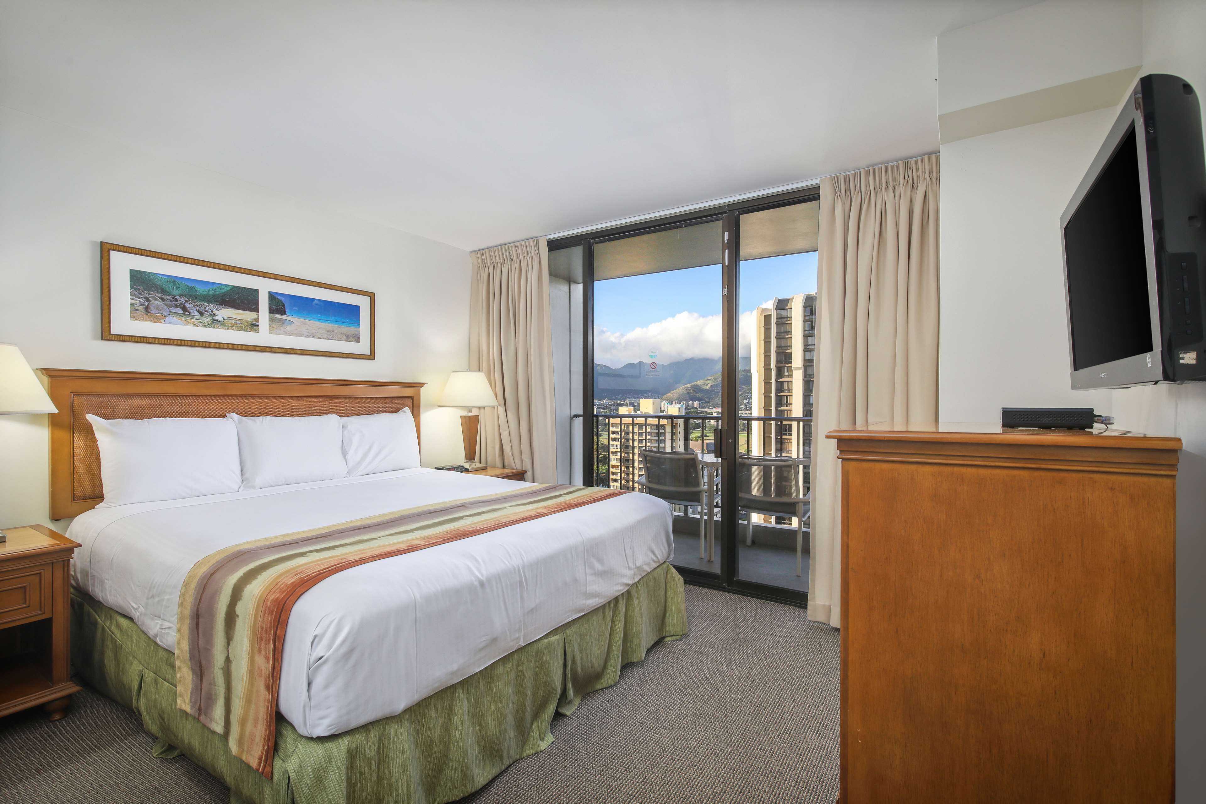 A hotel room with a large bed, two bedside tables with lamps, a wooden desk, a flat-screen TV, and a balcony with a city view.