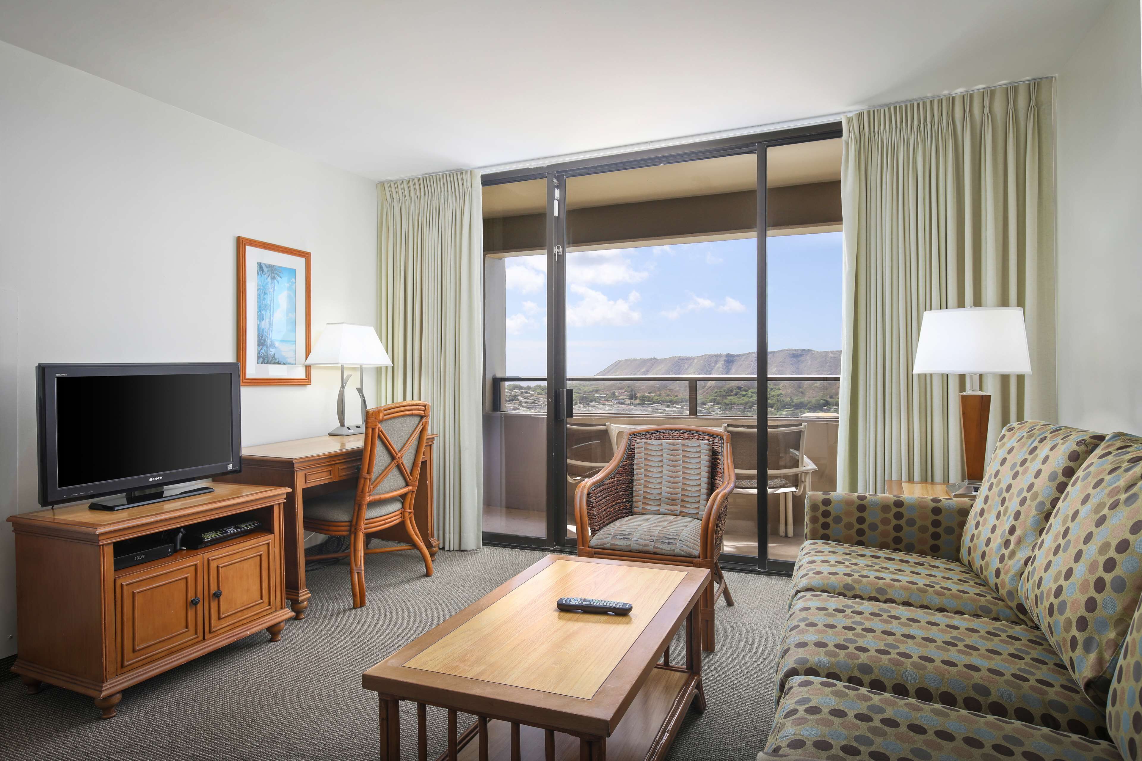 Habitación de hotel con televisor, mesa de centro de madera, sofá, sillón y balcón con vistas.