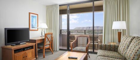 Habitación de hotel con televisor, mesa de centro de madera, sofá, sillón y balcón con vistas.