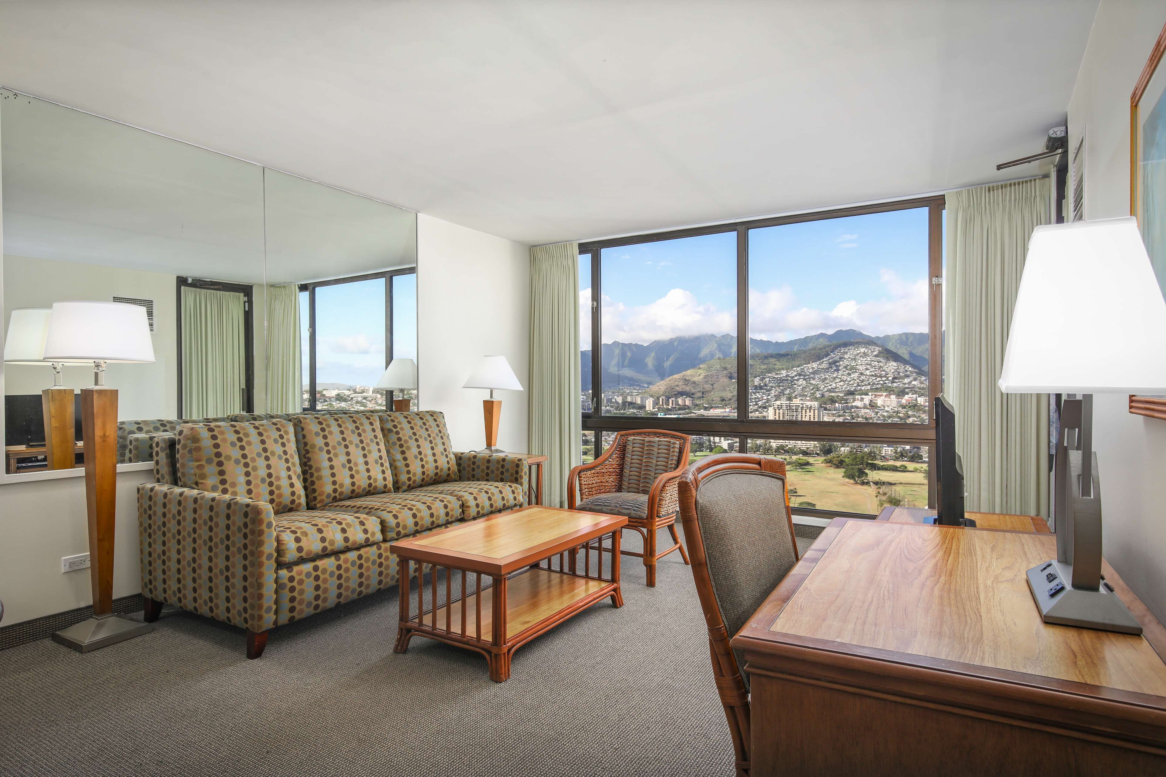A hotel room with a sofa, armchair, and a desk with a lamp. There is a large window offering a view of a city and mountains.