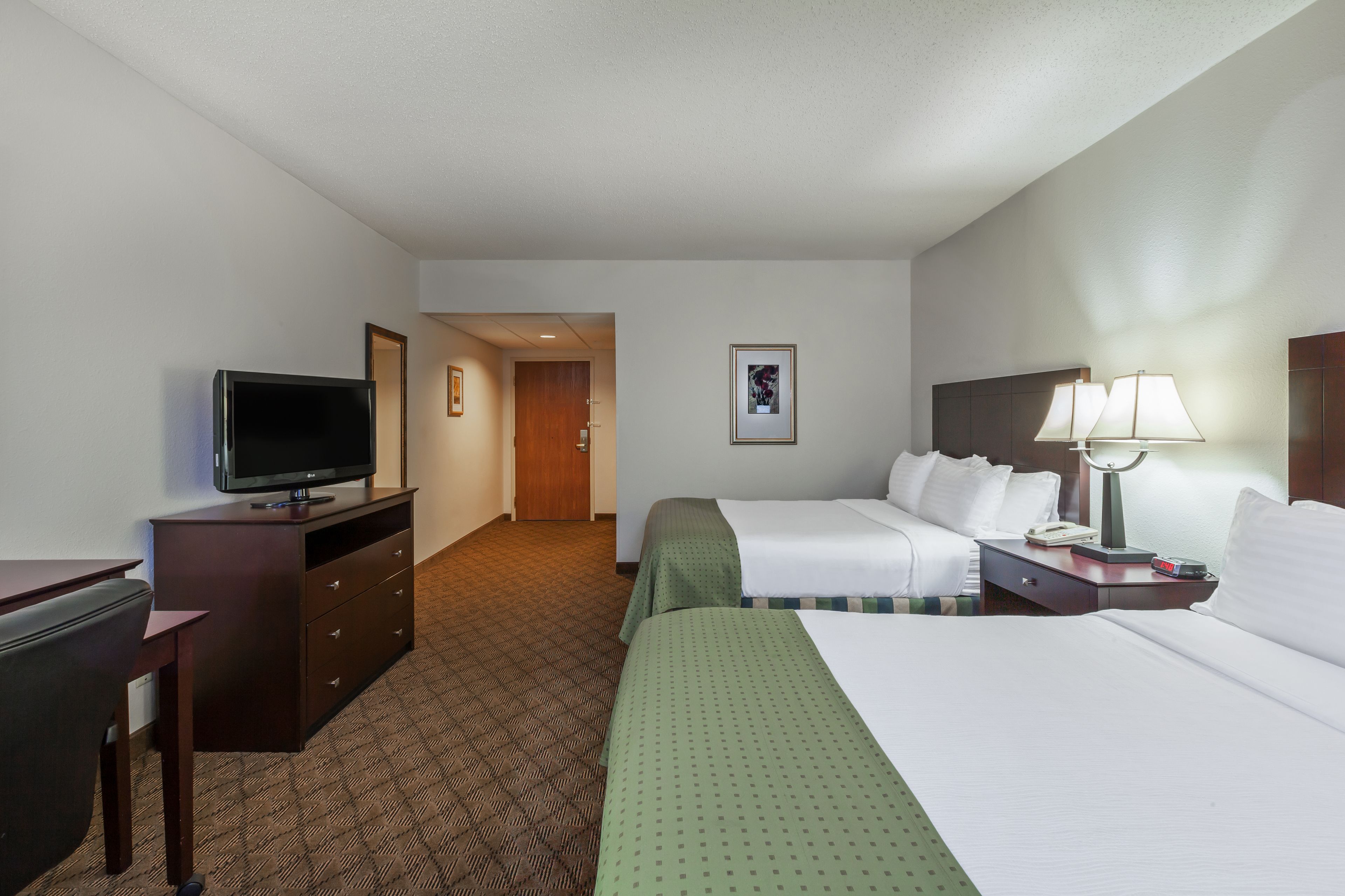 A hotel room with a bed, a desk with a television, a lamp, and a door leading to another room.