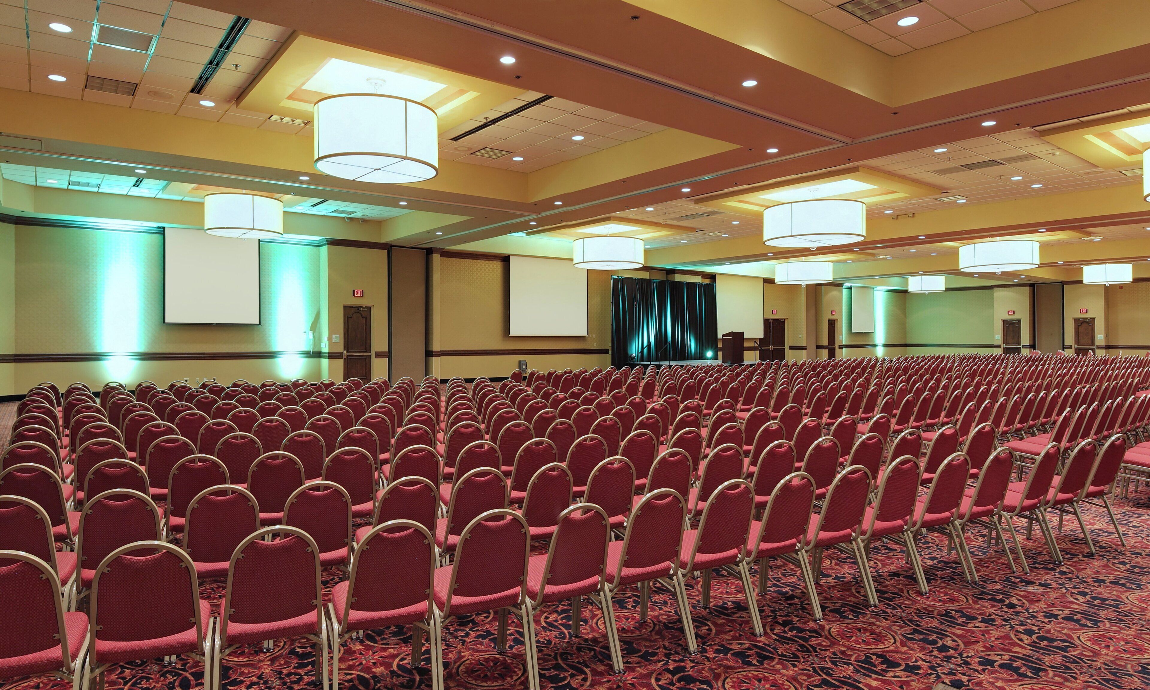 A conference room with rows of chairs, a stage, and a large screen.