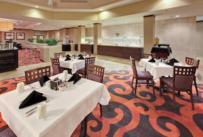 Uma sala de jantar de hotel com mesas preparadas para hóspedes, uma mesa de buffet e uma receção.