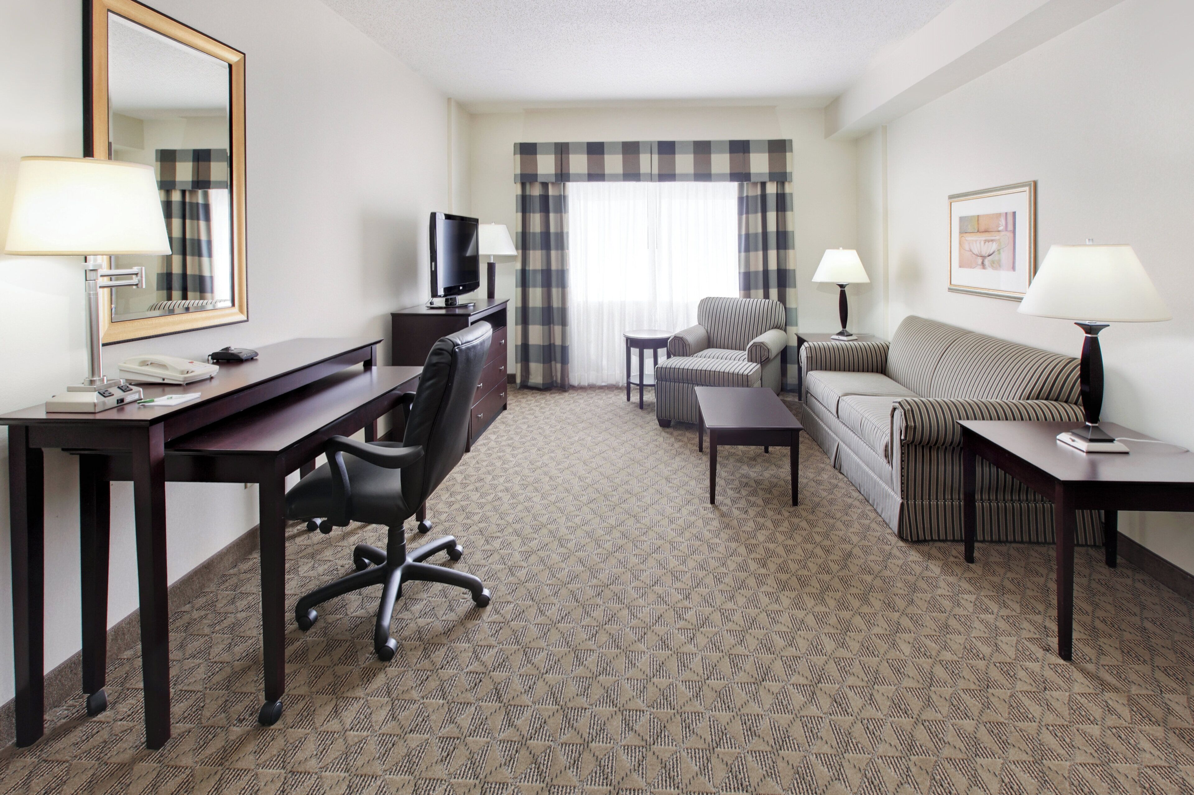 A hotel room with a desk, office chair, television, sofa, and two side tables with lamps.
