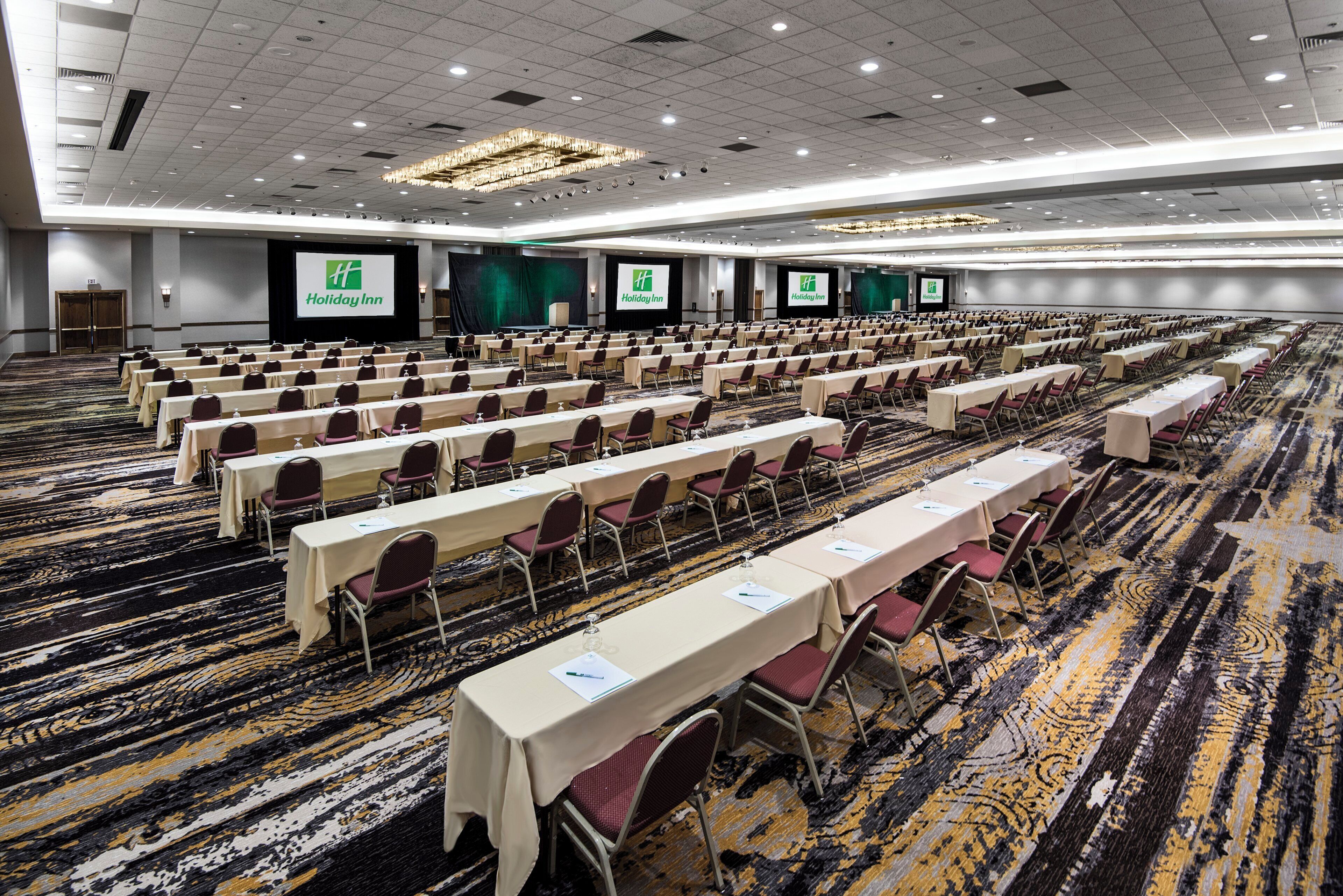Une grande salle de conférence avec des rangées de tables et de chaises, un sol recouvert de moquette et une scène sur laquelle est affiché le logo de Holiday Inn.