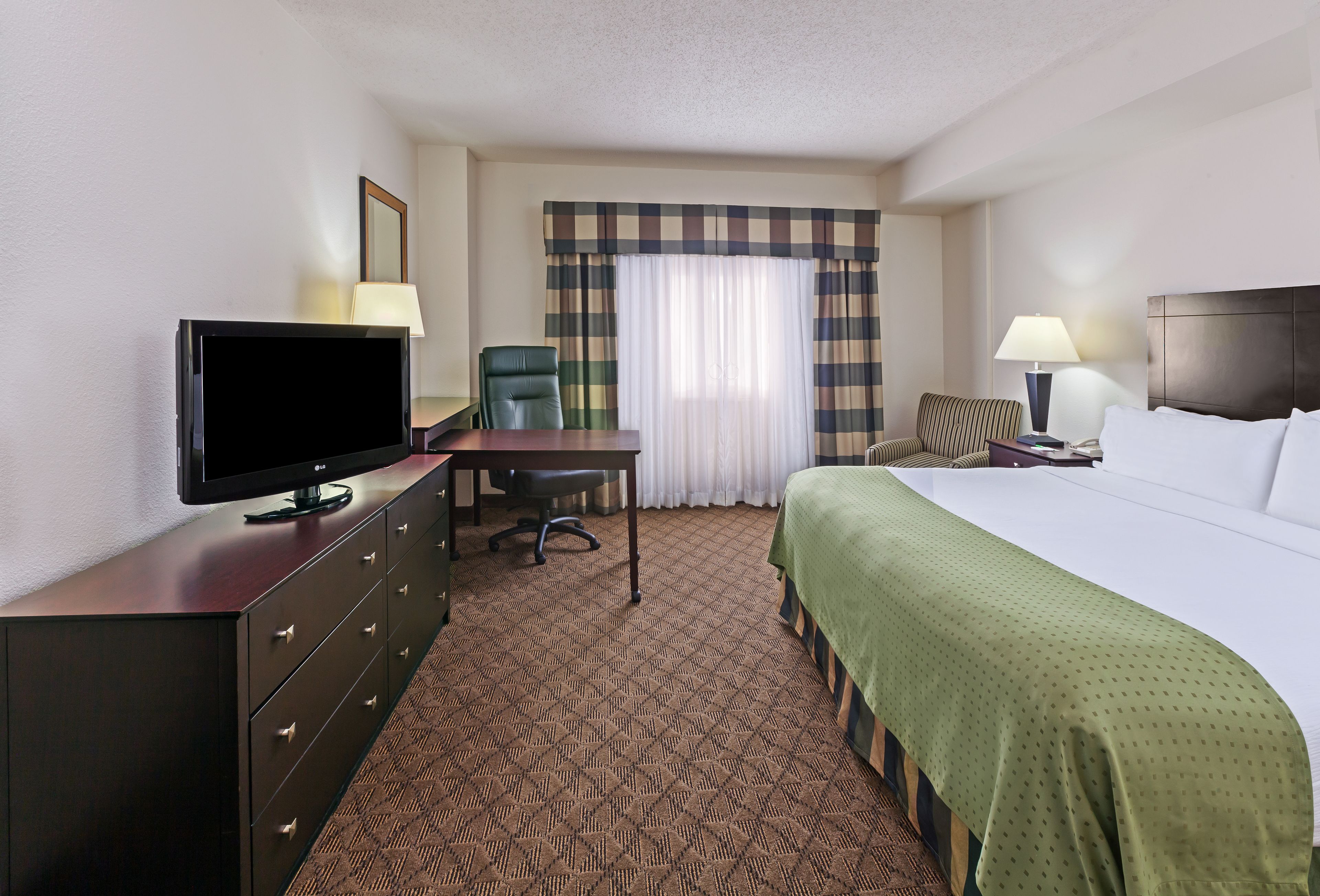 A hotel room with a bed, a desk with a television, a chair, a lamp, and a window with curtains.