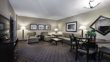 Una camera d'hotel con un'area salotto, che include una divano, due poltrone, un tavolino e un tavolo da pranzo con sedie. Alle pareti ci sono quadri incorniciati, una TV a schermo piatto montata sopra e un vaso con una pianta sul tavolo da pranzo.