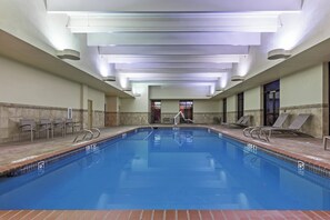Una amplia piscina interior con agua cristalina, rodeada de sillas de descanso y un suelo de baldosas.