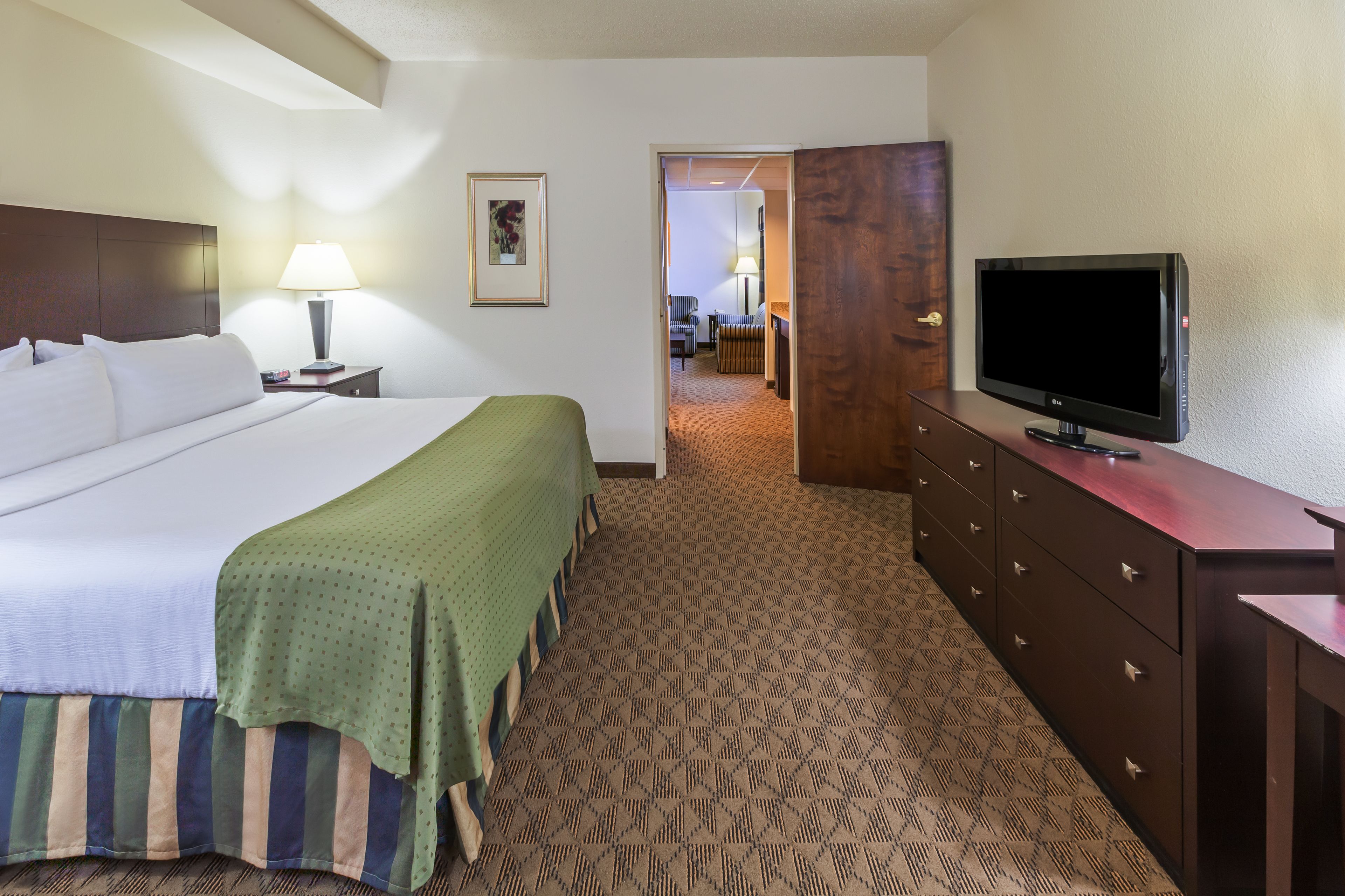 A hotel room with a large bed, a television on a dresser, a lamp, and a door leading to another room.