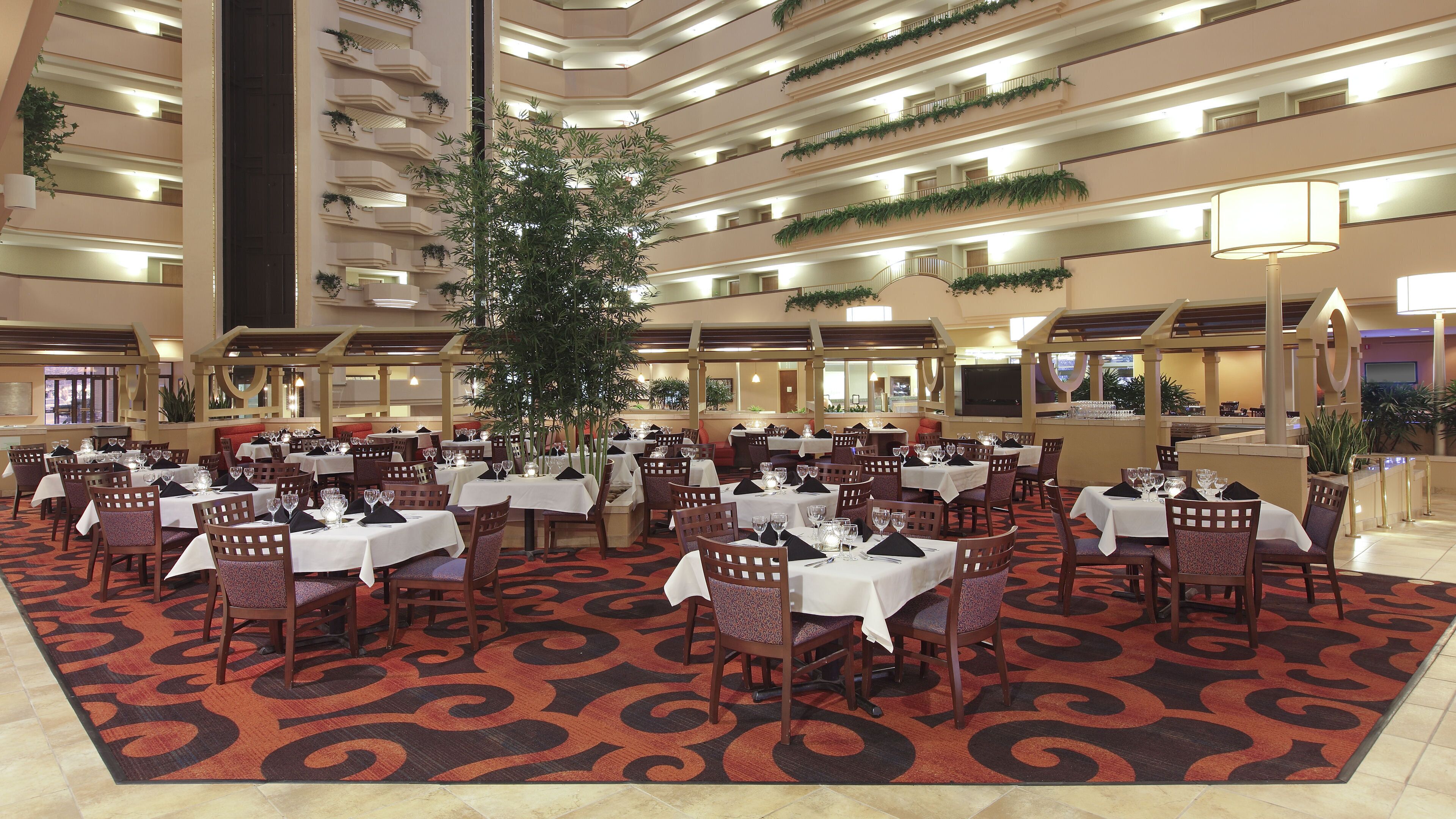 Ein Hotelrestaurant mit mehreren Tischen und Stühlen, einer großen Pflanze in der Mitte und einem gemusterten Teppich.