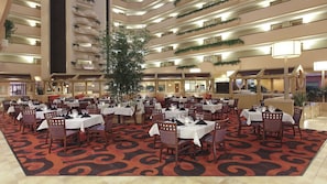 A hotel dining area with multiple tables and chairs, a large central plant, and a patterned carpet.