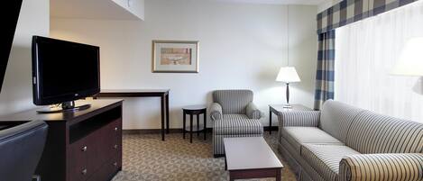A hotel room with a flat-screen TV, a framed picture on the wall, a striped armchair, a sofa, a small table, and a lamp.