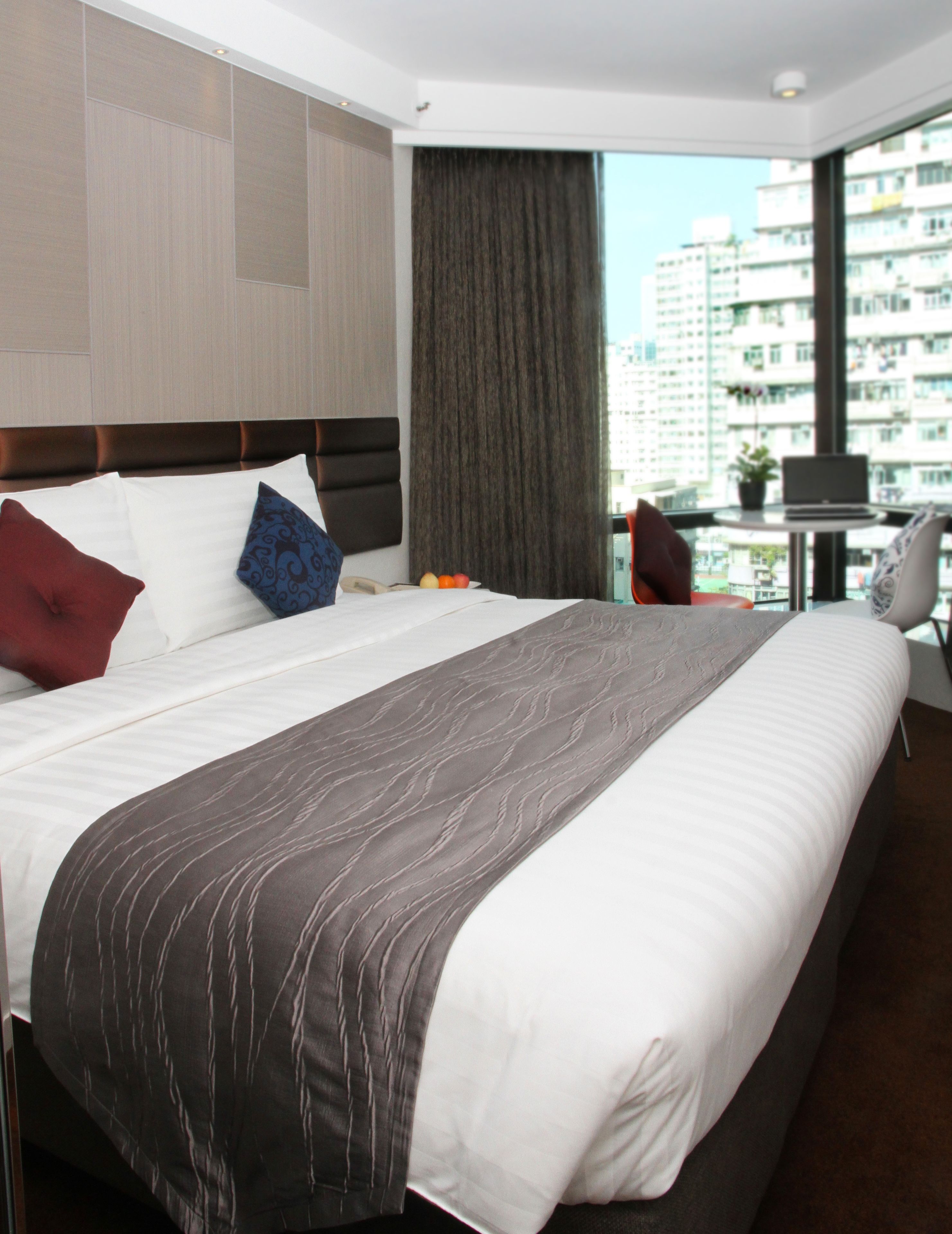 A hotel room with a large bed, a view of the city through the window, and a small table with a laptop.