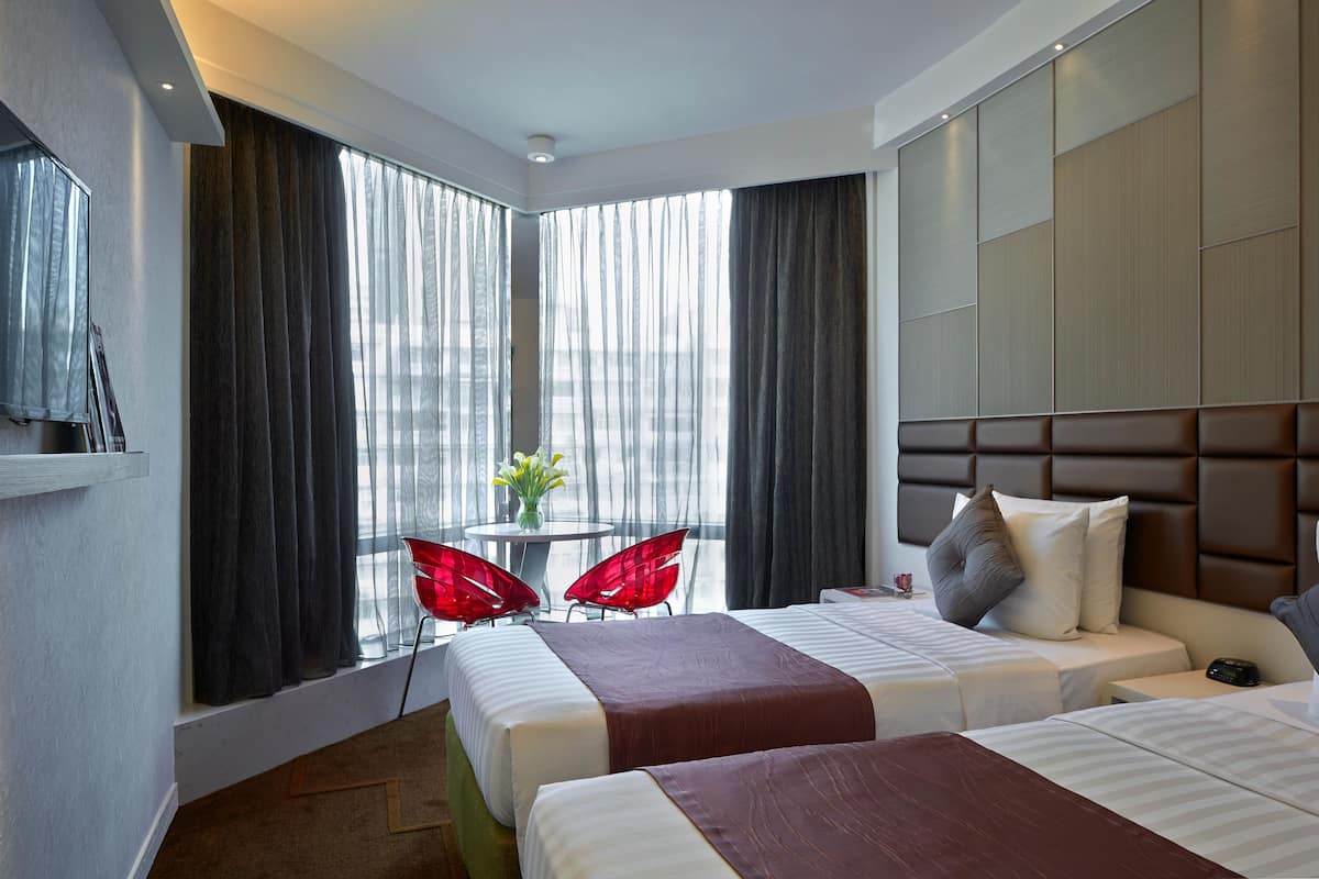 A hotel room with two beds, a TV, a small table with chairs, and a view of the city through large windows.