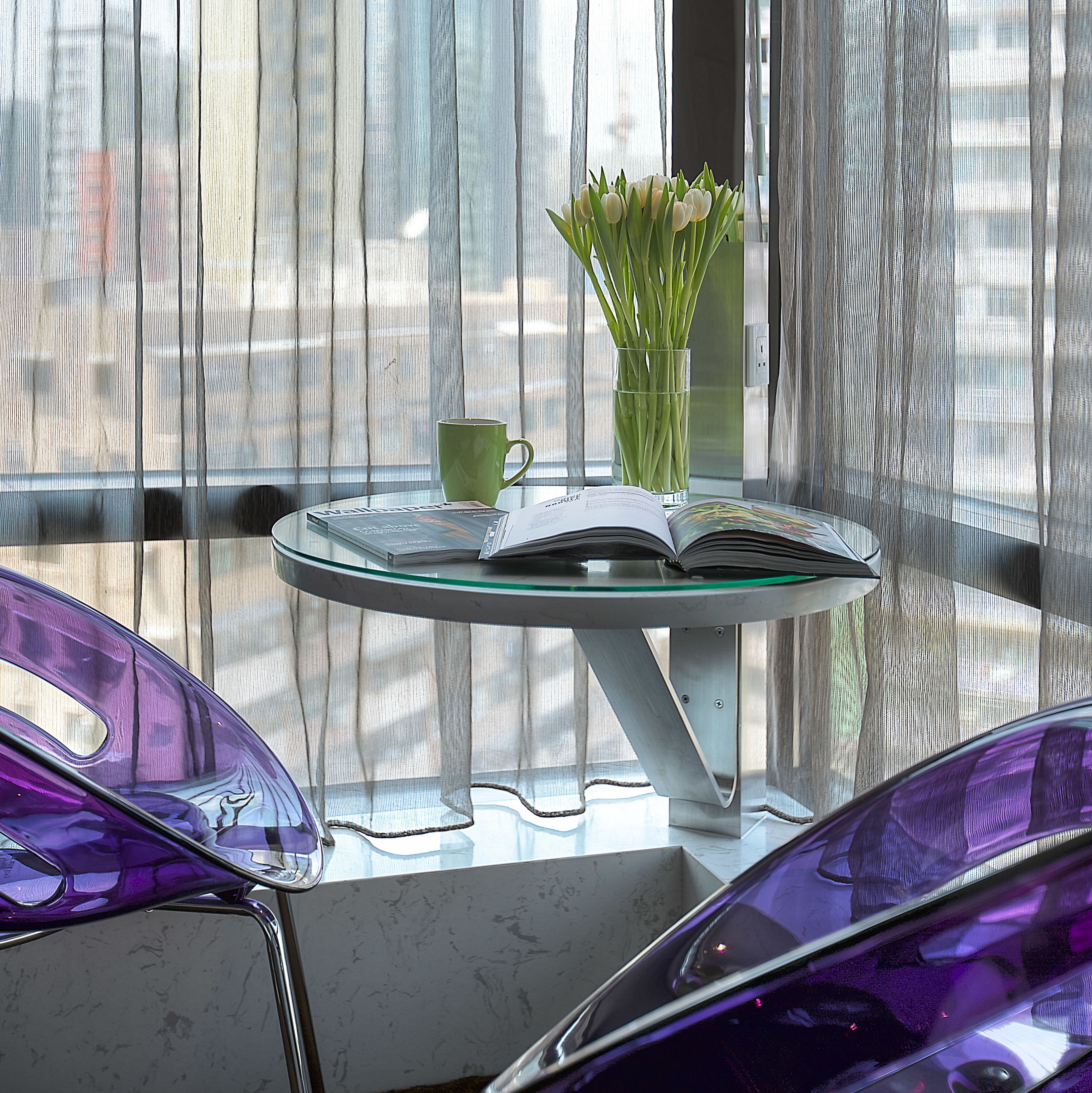 A glass table with a book and a green mug, a vase with green plants, and purple chairs in a room with sheer curtains.