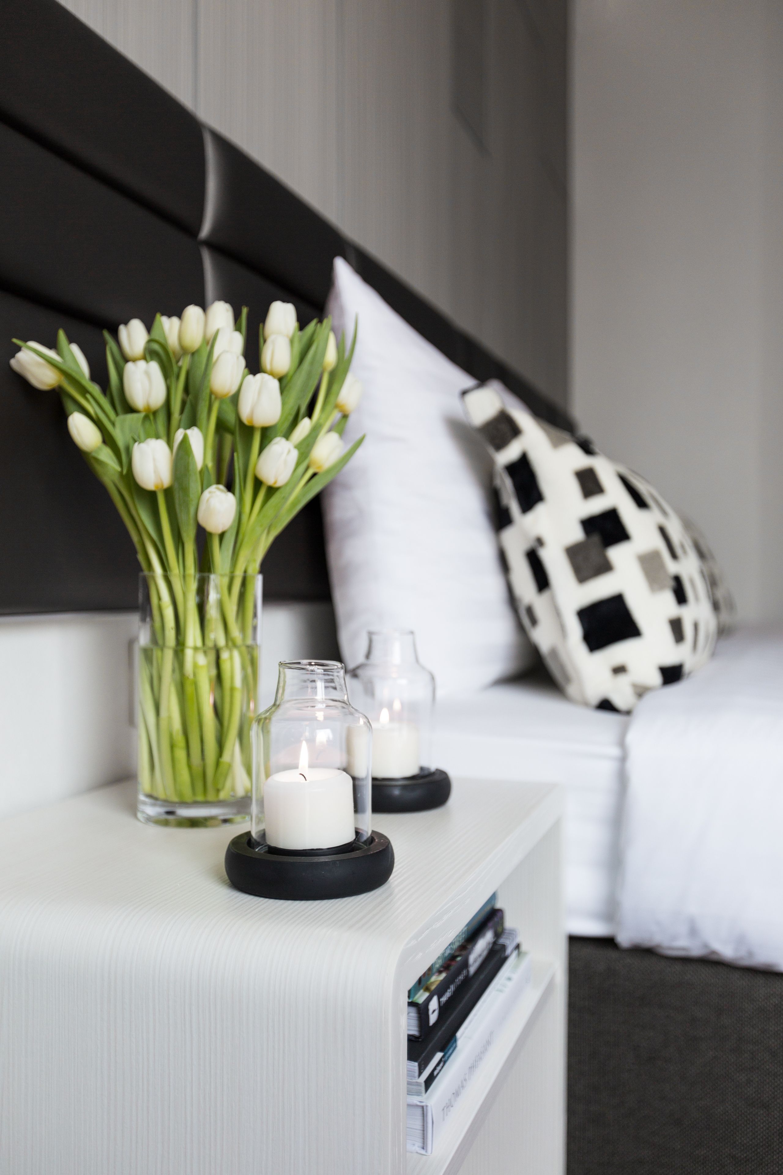 A neatly arranged bedside table with a vase of white tulips, two lit candles, and a few books.
