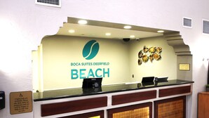 Un mostrador de recepción con un cartel que dice "Boca Suites Deerfield Beach" y una computadora Dell. Hay dos mostradores, un cartel sobre accesibilidad para sillas de ruedas y un elemento decorativo en la pared.