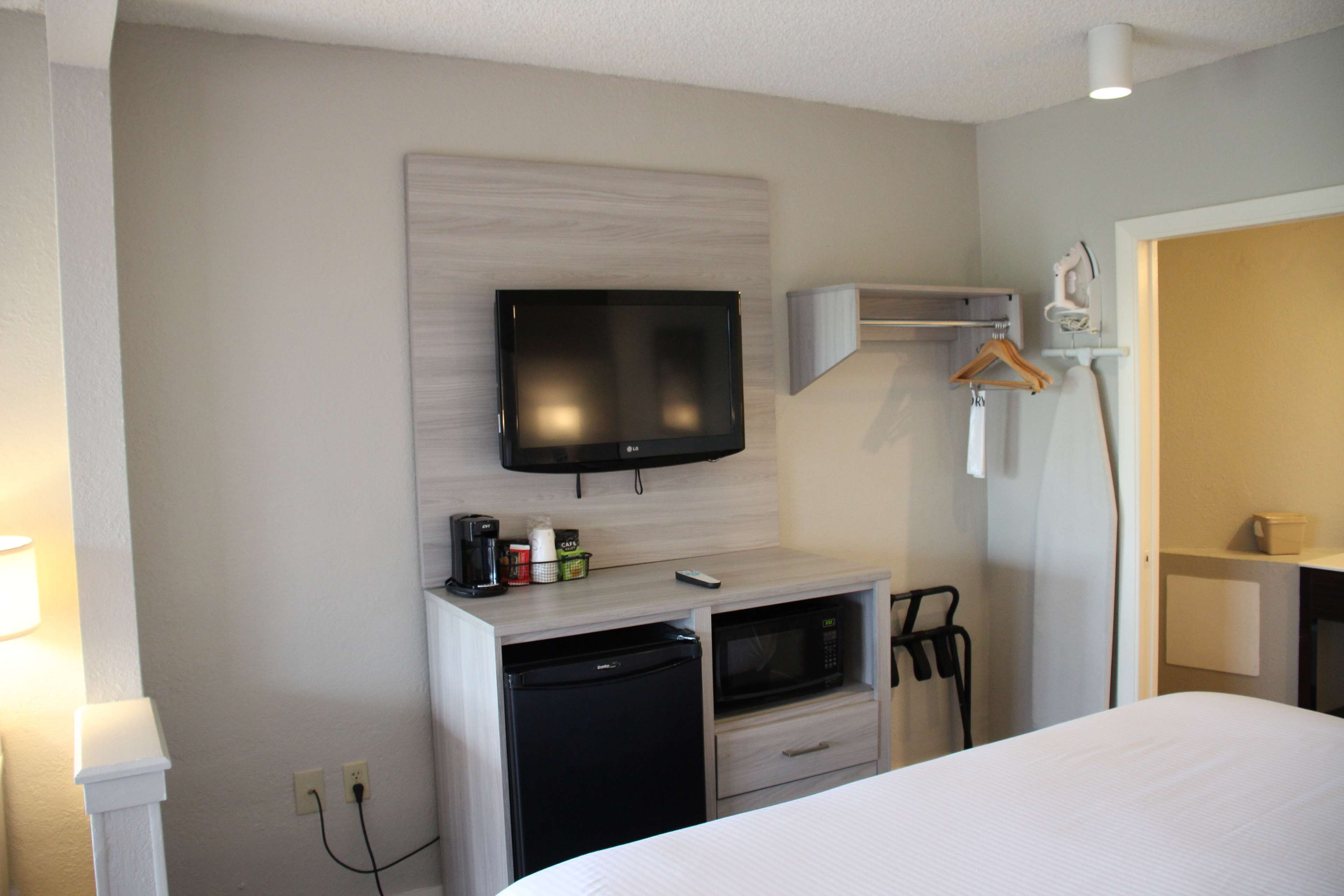 A hotel room with a bed, a TV mounted on the wall, a microwave, a small refrigerator, a desk with a chair, and a hanging coat rack.