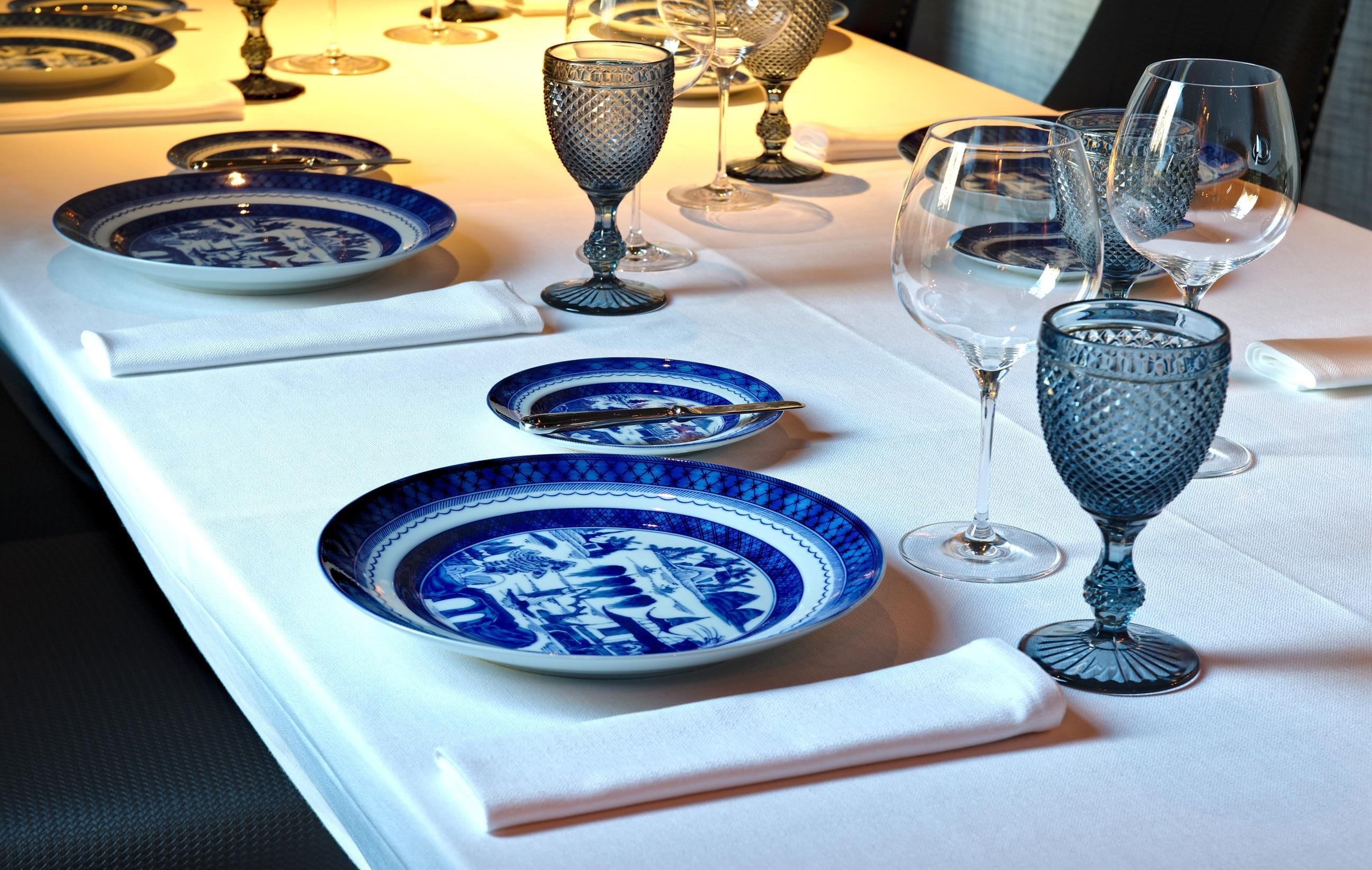 A table set with blue and white patterned plates, clear wine glasses, and a white tablecloth.