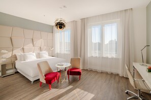 Una camera d'albergo moderna con un letto grande, due poltrone rosse, un piccolo tavolino rotondo e una scrivania con una lampada.