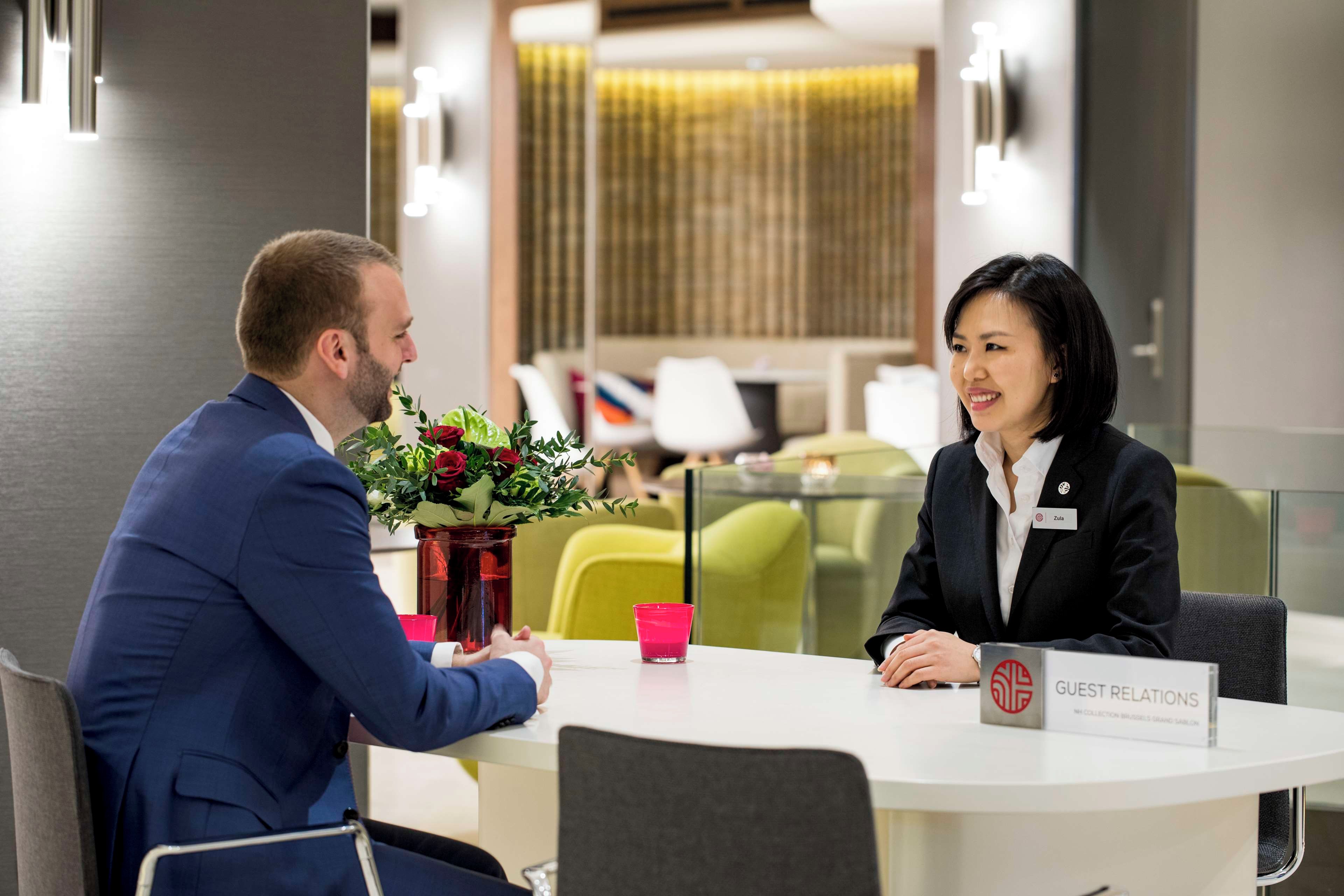 A professional setting with a man and a woman seated at a table, a name tag, and a 'GUEST RELATIONS' sign.