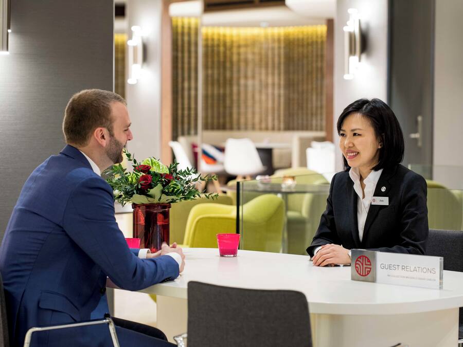 A professional setting with a man and a woman seated at a table, a name tag, and a 'GUEST RELATIONS' sign.