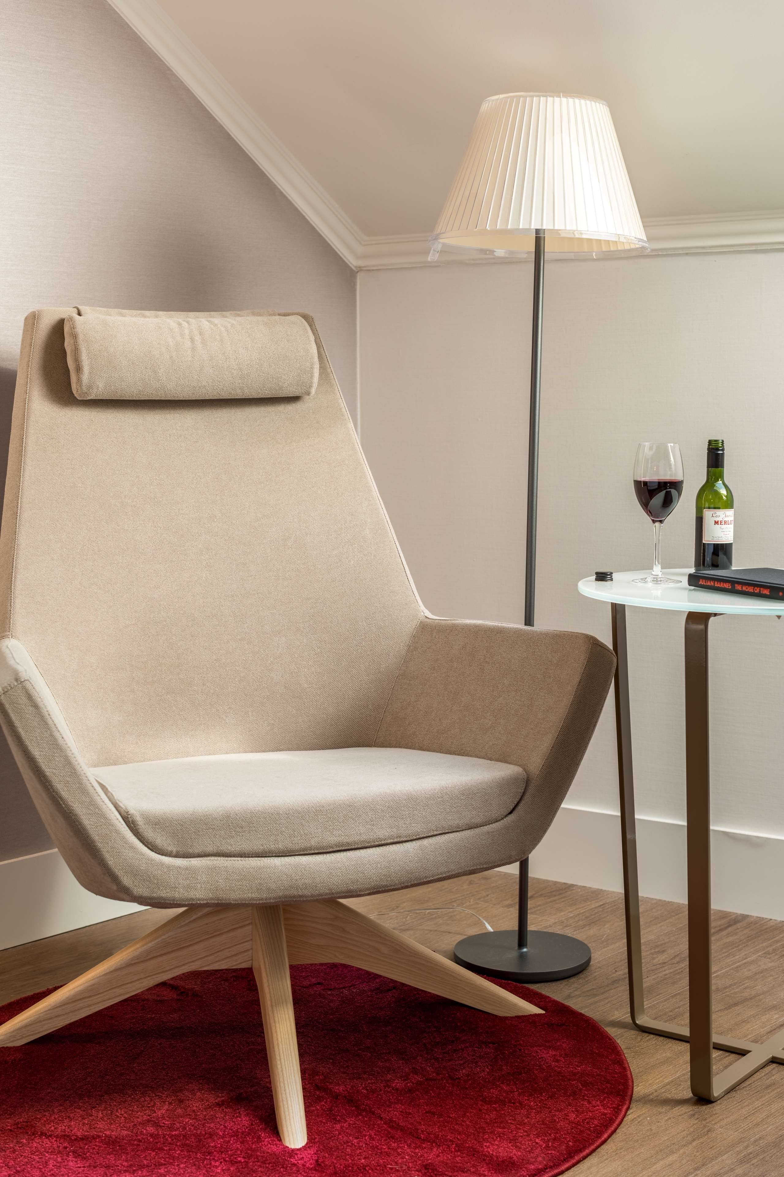 A modern chair with a high backrest, a round table with a lamp, and a bottle of wine.