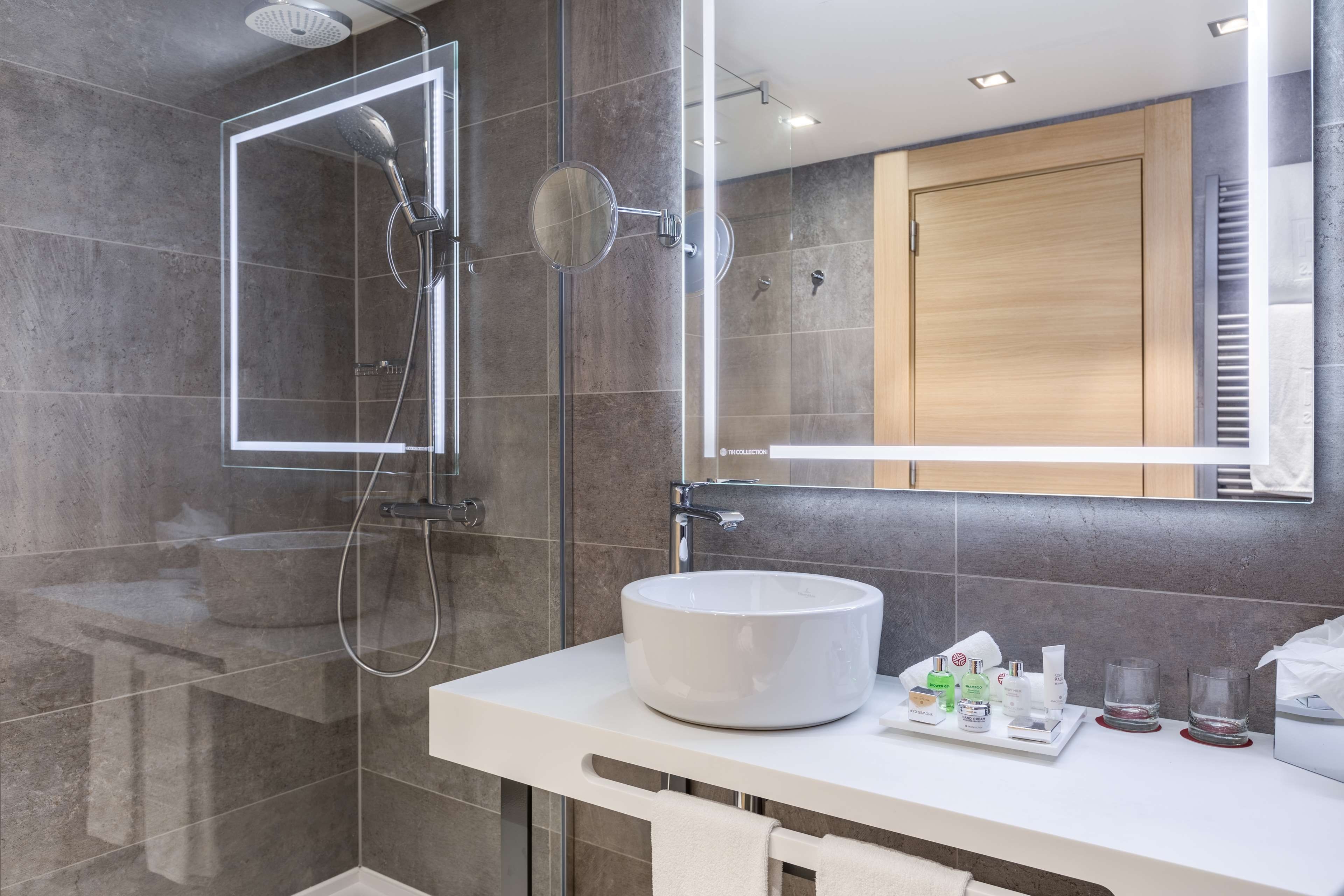 Un baño moderno con ducha de vidrio, lavamanos blanco e iluminación integrada en el espejo.