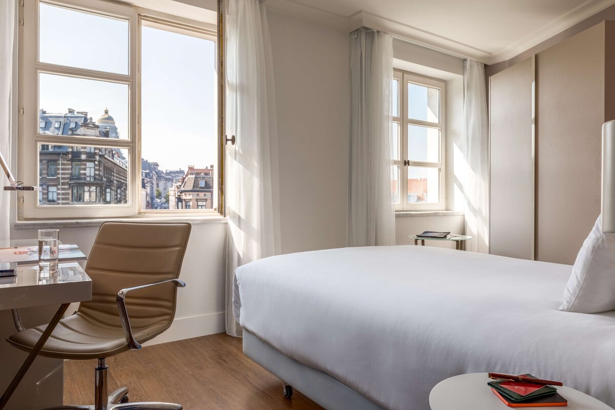 Camera d'albergo con un letto, una scrivania con una sedia, un tavolino e vista sui palazzi dalla finestra.