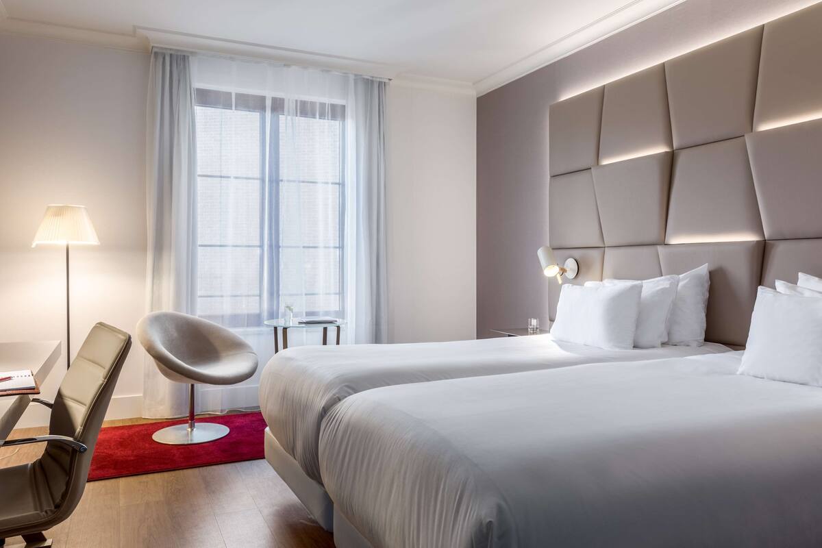 A hotel room with two beds, a desk, a chair, a lamp, and a window with curtains.