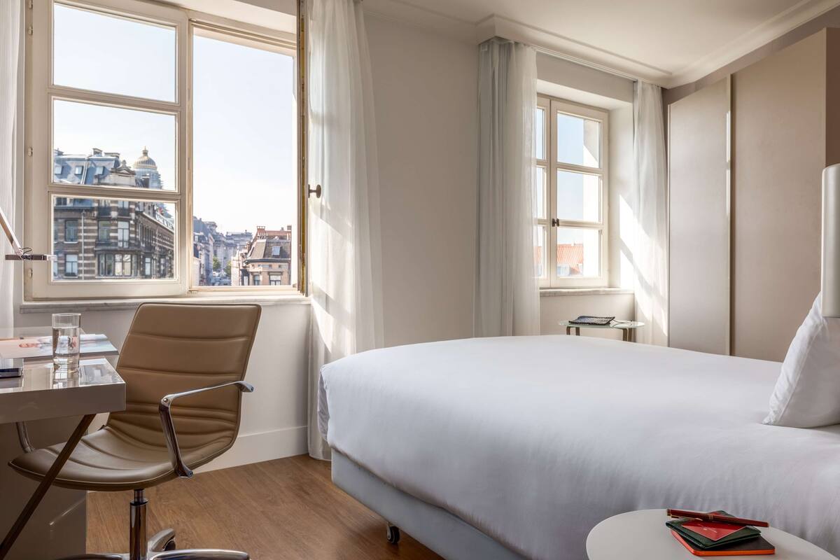 A hotel room with a bed, a desk with a chair, a small table, and a view of buildings through the window.