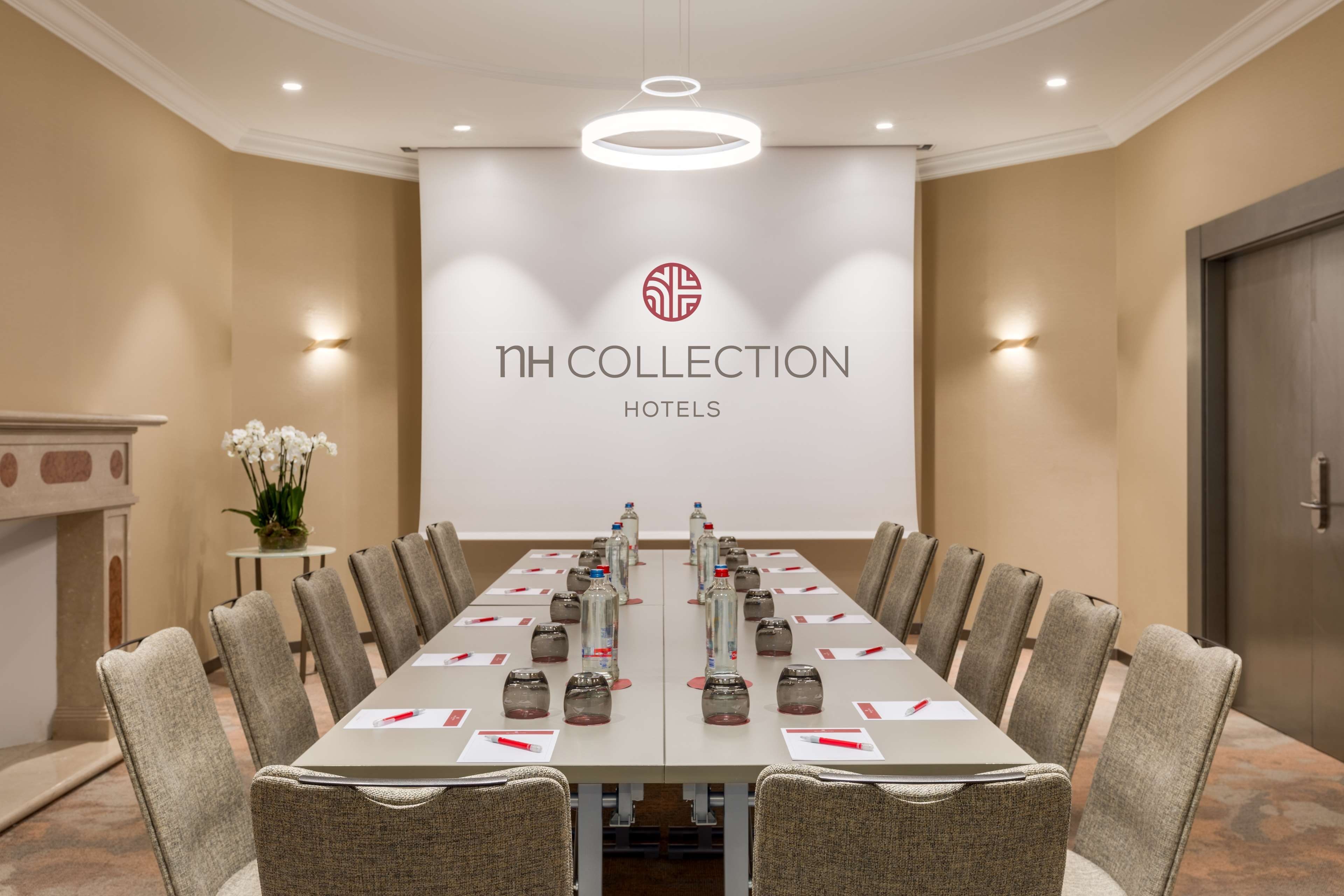 A conference room with a long table, chairs, water bottles, and a projector screen displaying 'NH COLLECTION HOTELS'.