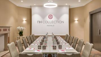 A conference room with a long table, chairs, water bottles, and a projector screen displaying 'NH COLLECTION HOTELS'.