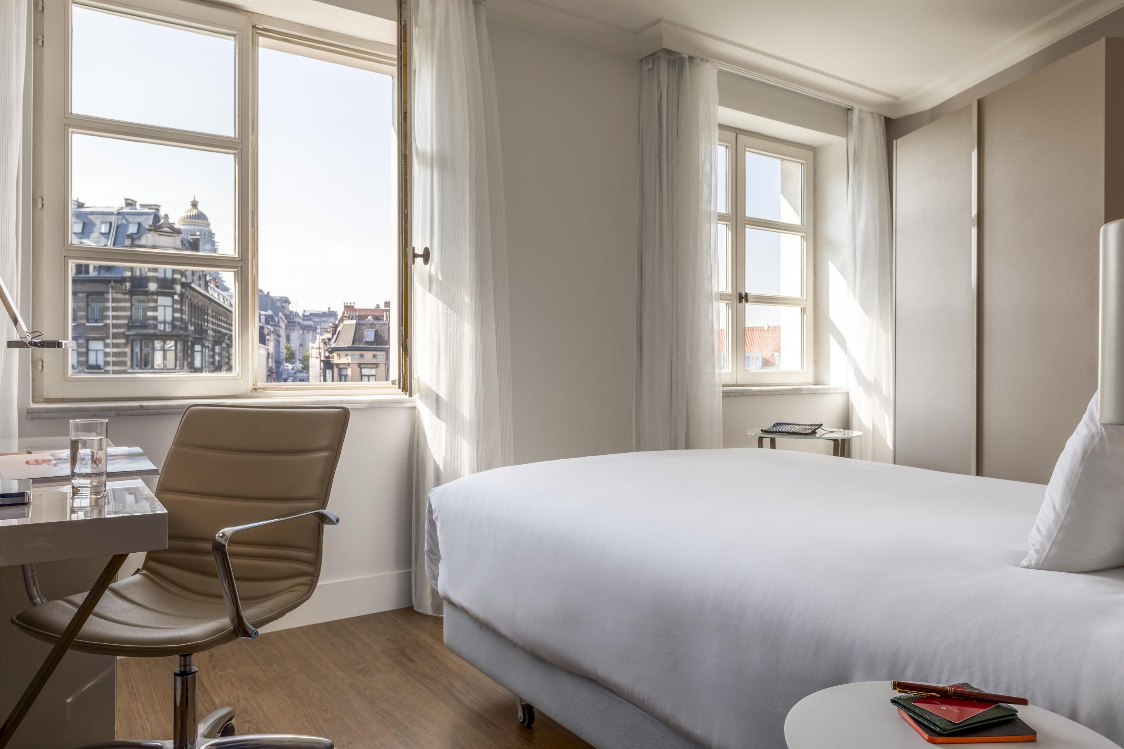 A hotel room with a bed, a desk, a chair, and a view of the cityscape through large windows.