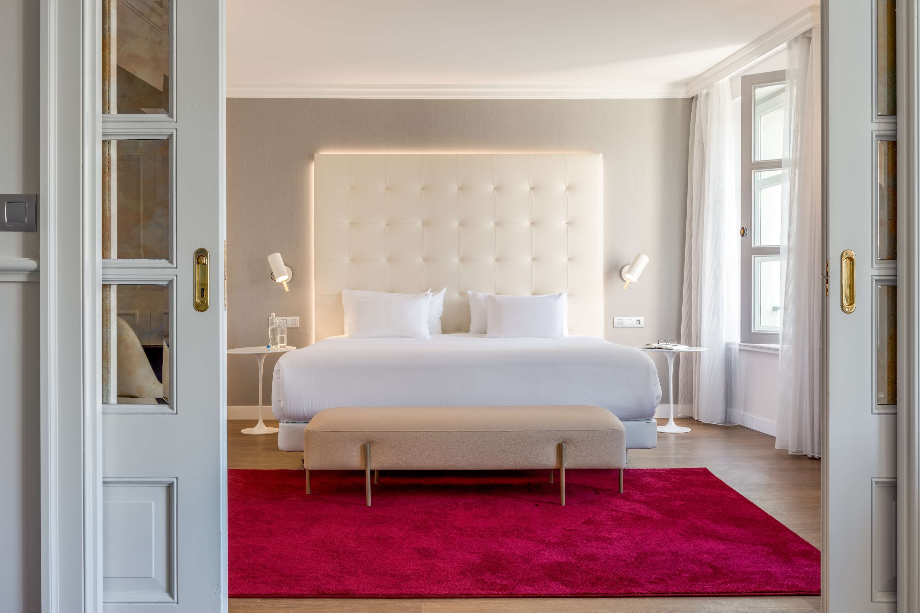 A bedroom with a large bed, bedside tables, a bench, and a red rug.