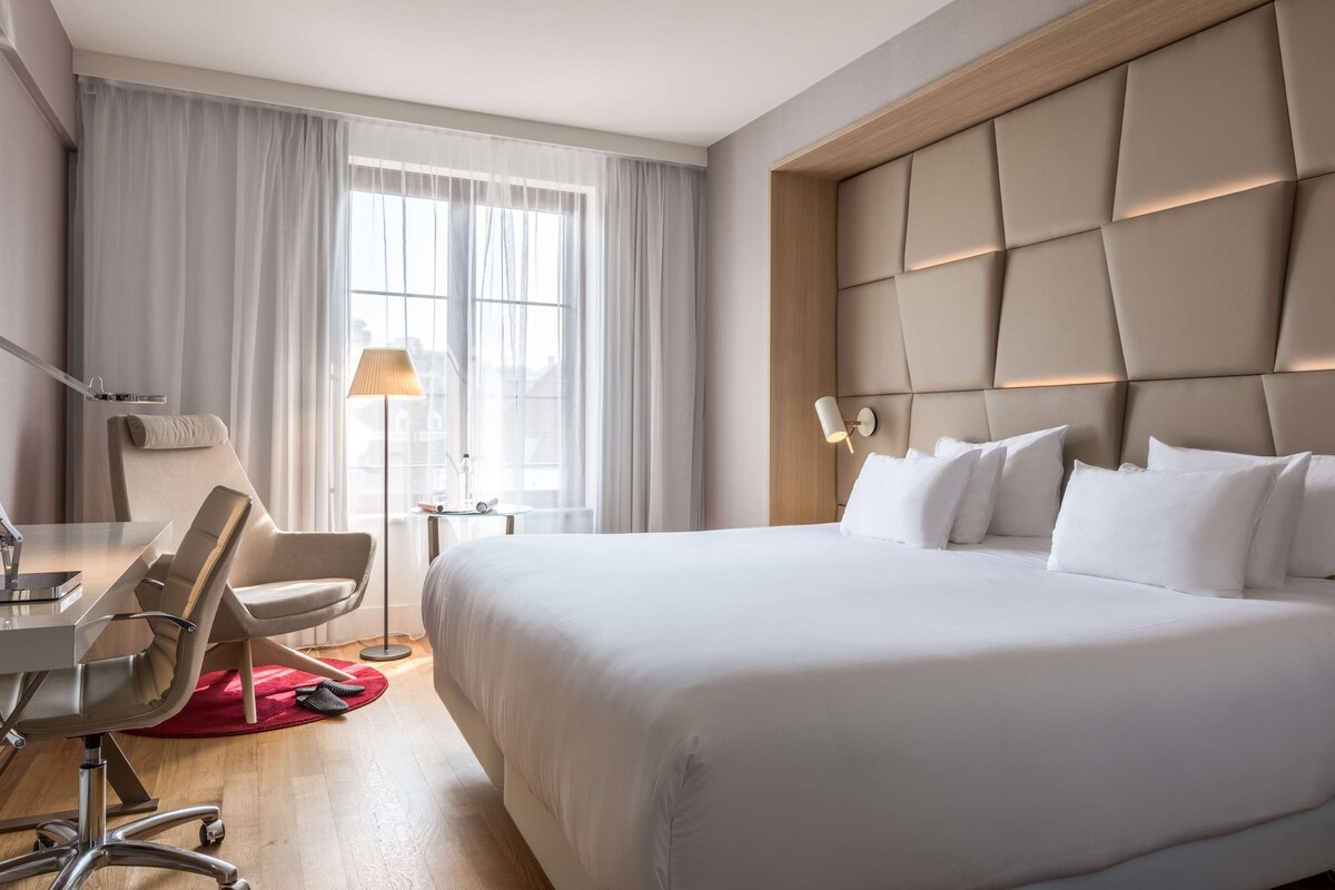 Una camera d'albergo con un letto, una scrivania, una sedia, una lampada e una finestra con tende.