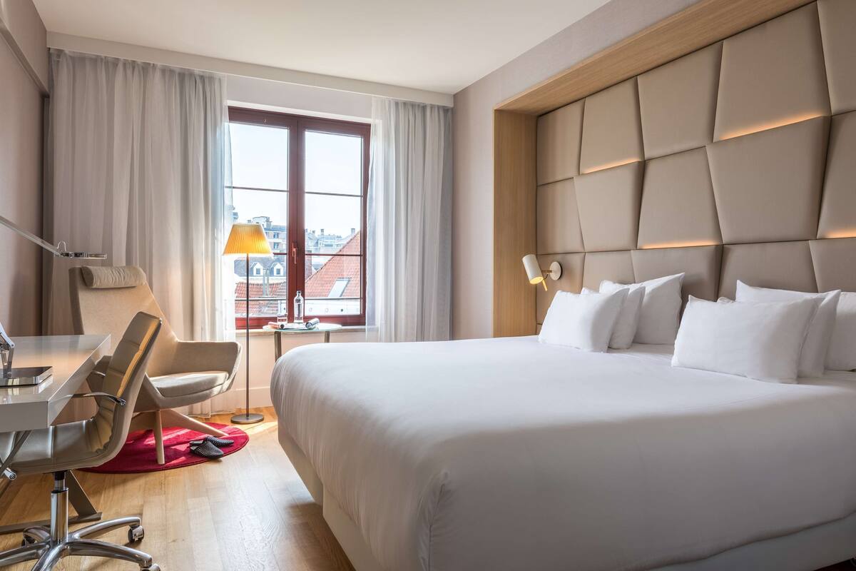 A hotel room with a large bed, a desk with a chair, a lamp, and a view of buildings through a window.