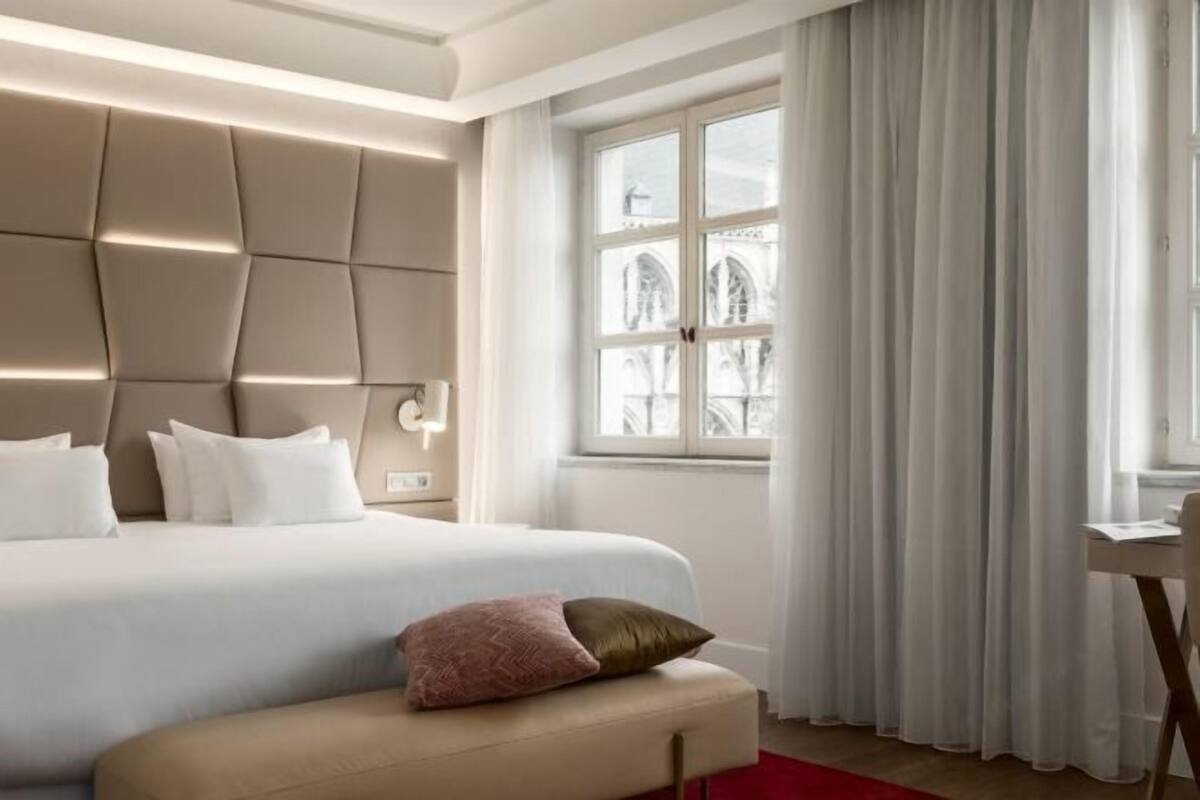 A modern hotel room with a large bed, a cushioned bench, a desk, and a window with curtains.