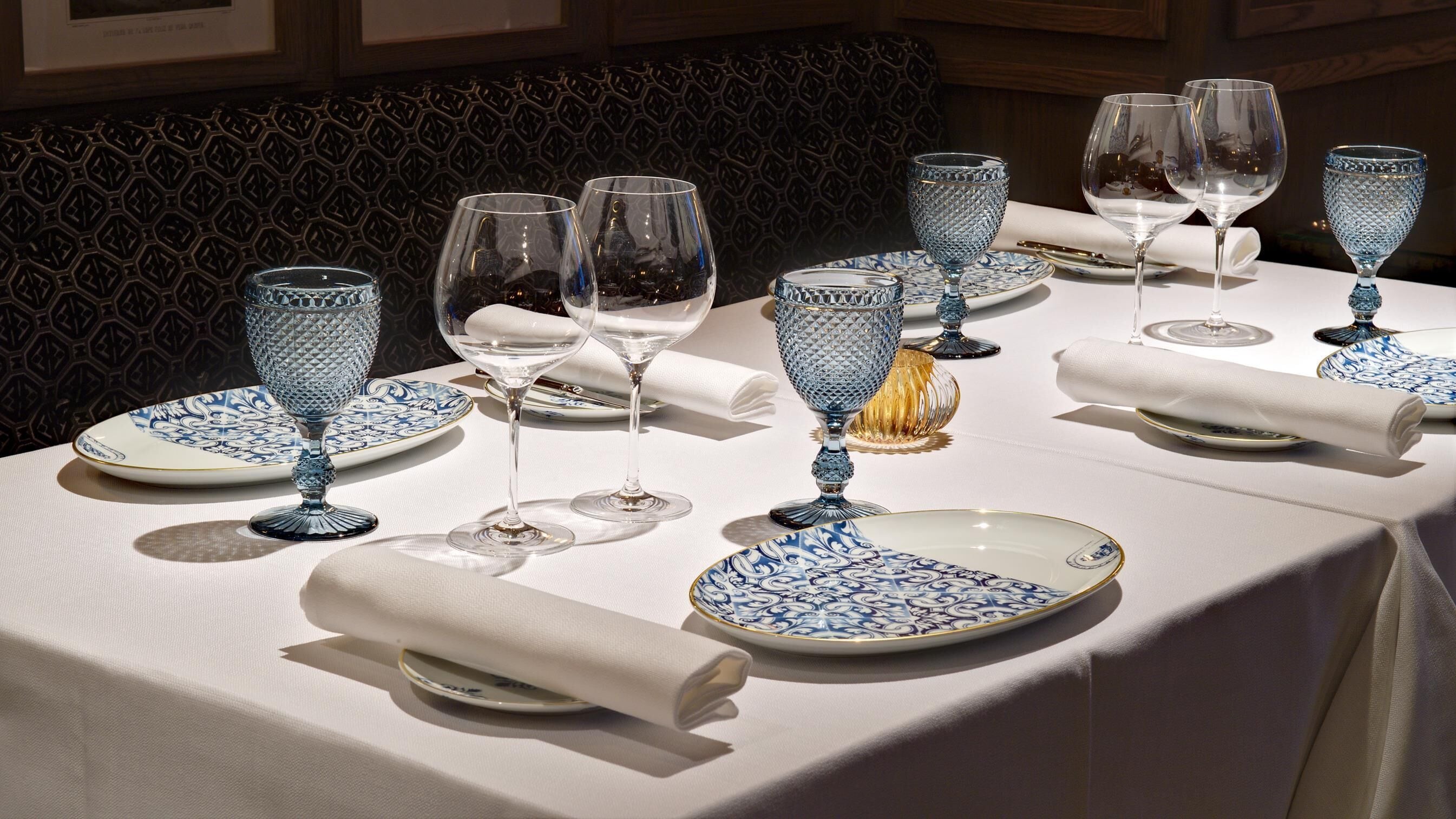 Una mesa dispuesta con platos azules y blancos, copas de cristal transparentes y un candelabro dorado.