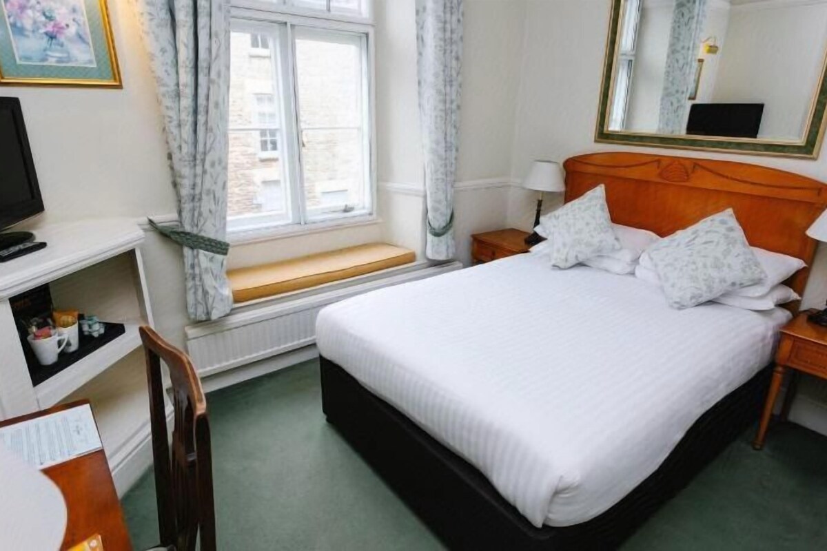 A hotel room with a bed, bedside tables, a desk with a chair, a television, and a window with curtains.